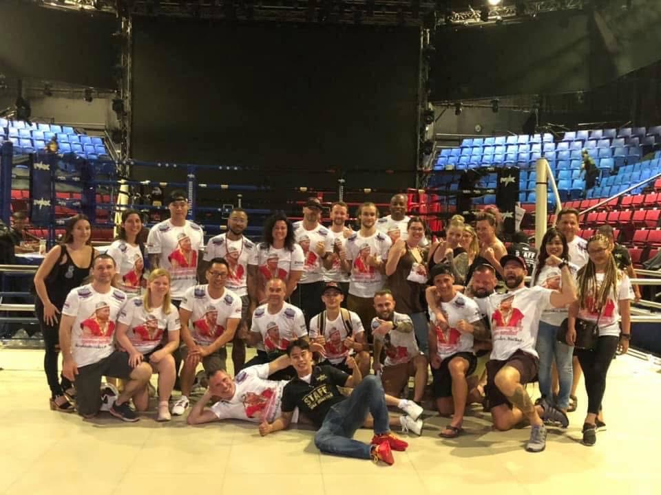 Group of people in a boxing ring, some wearing matching shirts, in a sports arena with seating around.