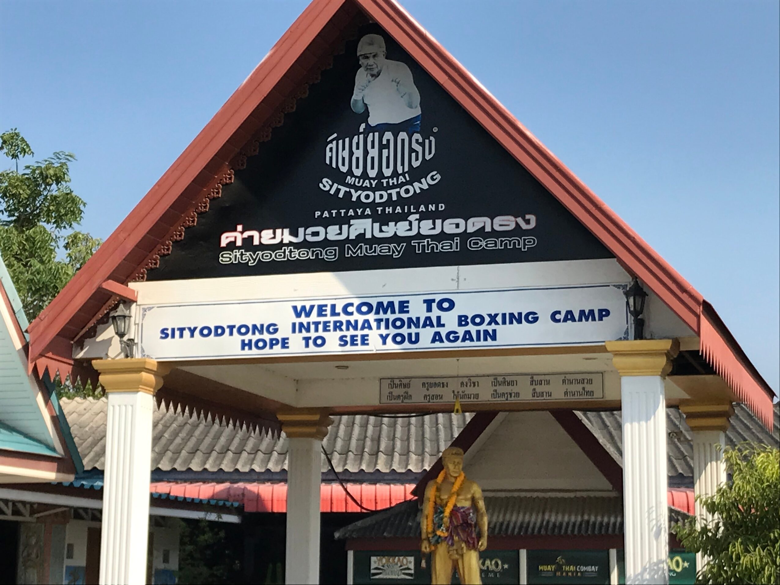 Entrance of Sityodtong International Boxing Camp with a welcome sign, columns, and a building with a triangular roof, blue sky.