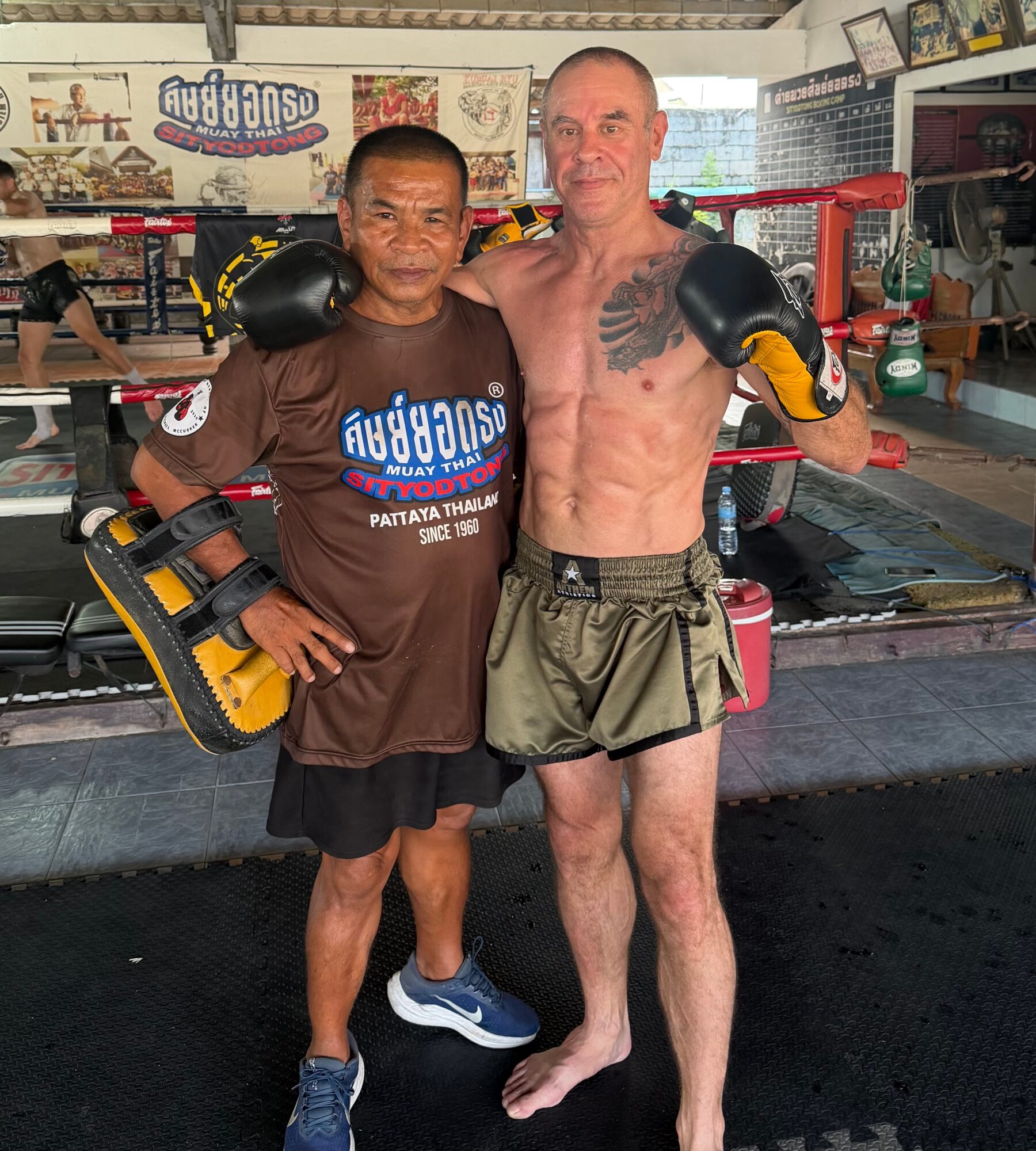 Two men in a boxing gym, one with boxing gloves and shorts, the other with boxing gloves and a T-shirt, smiling.