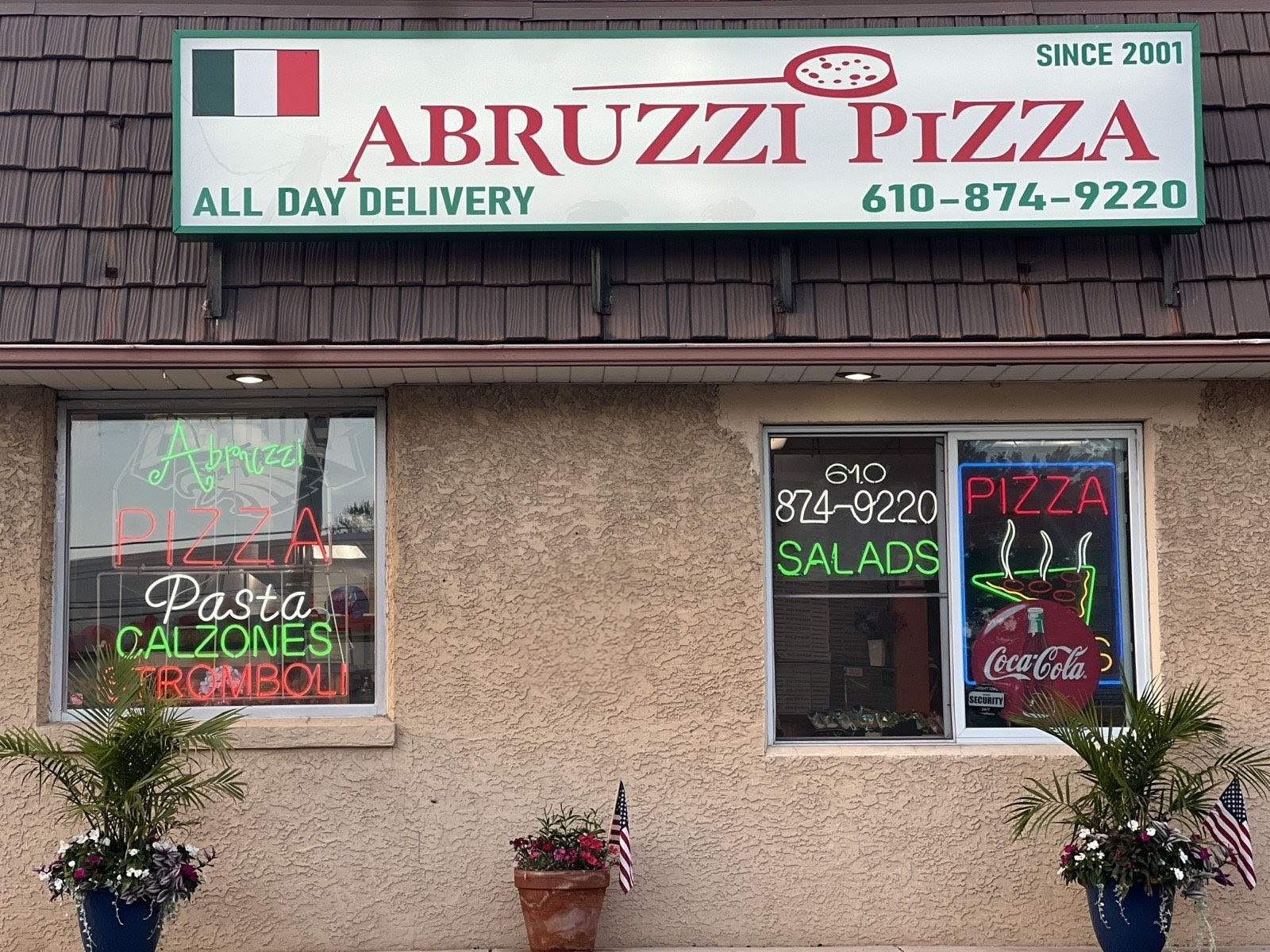 Pizza shop with signs for salads, pasta, calzones, and delivery, decorated with plants and an American flag, window displays neon signs.