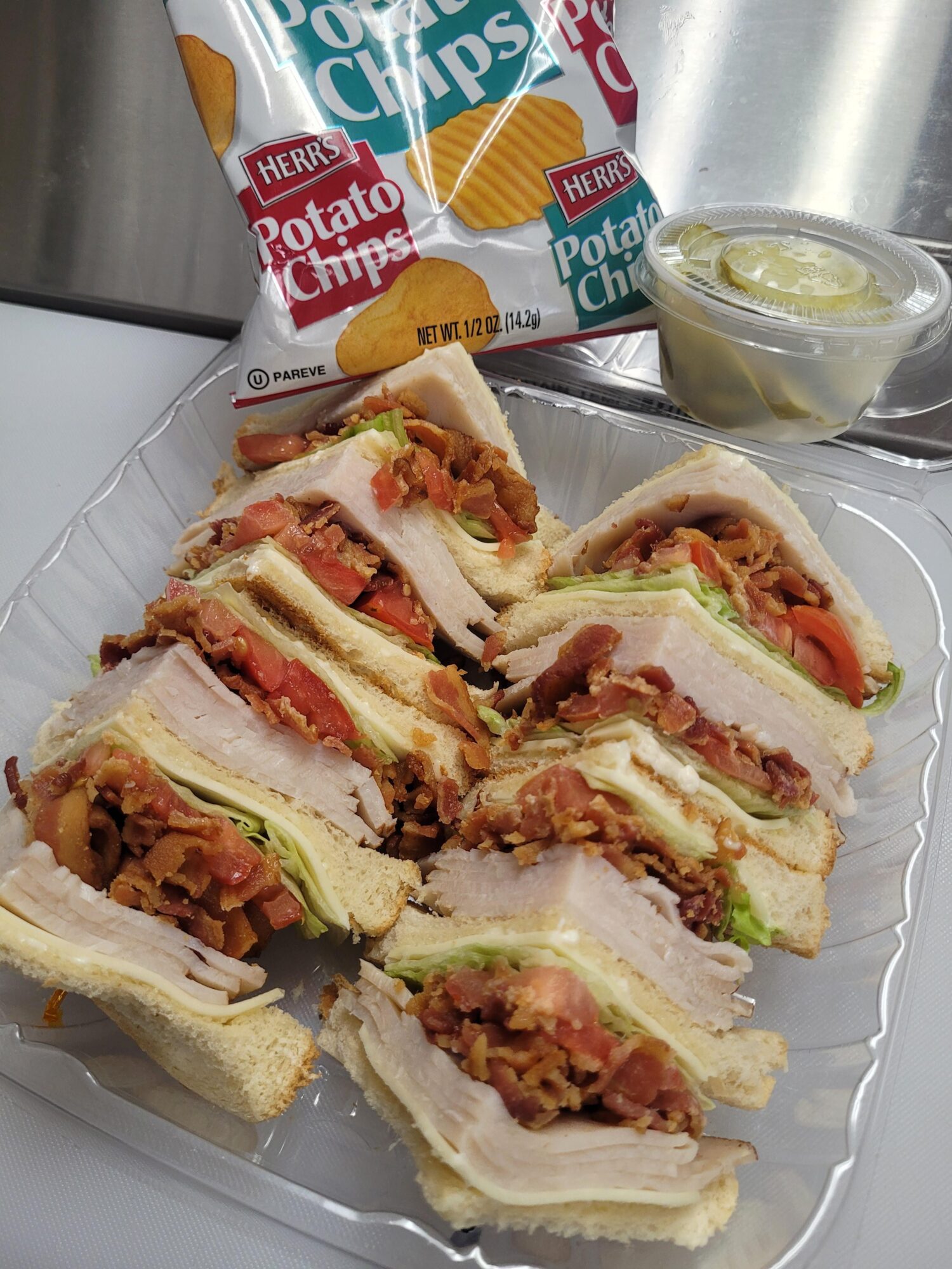 Plate with six sandwich halves filled with meat, lettuce, and tomato, alongside a small bowl of sauce and potato chips in the background.
