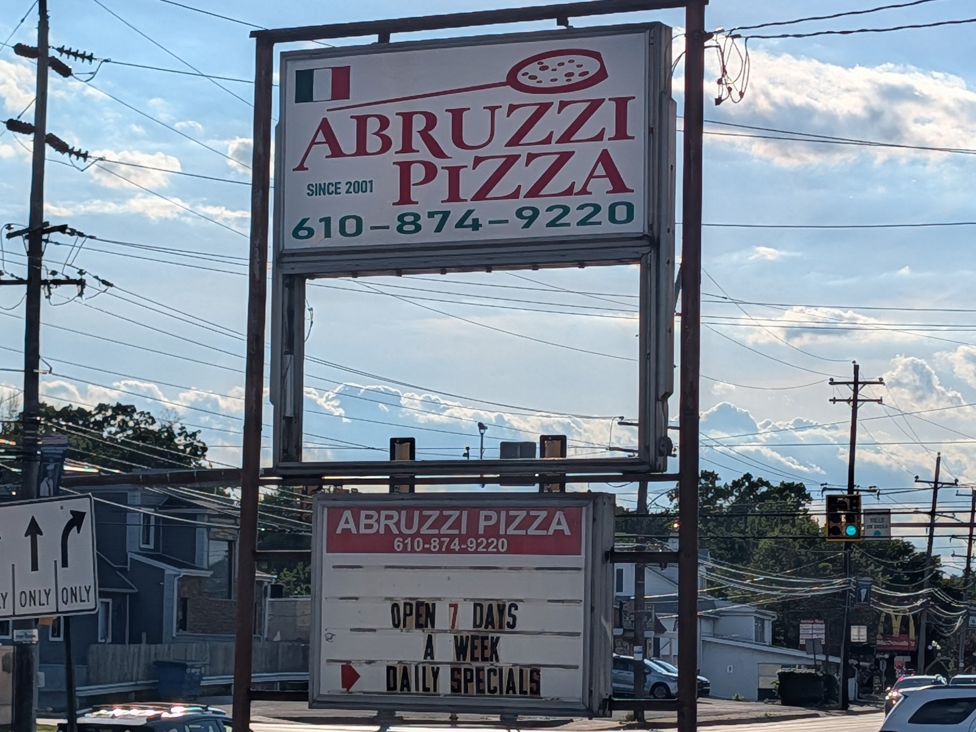 Sign for Abruzzi Pizza with phone number, open 7 days, weekly and daily specials, against a background of power lines and sky.