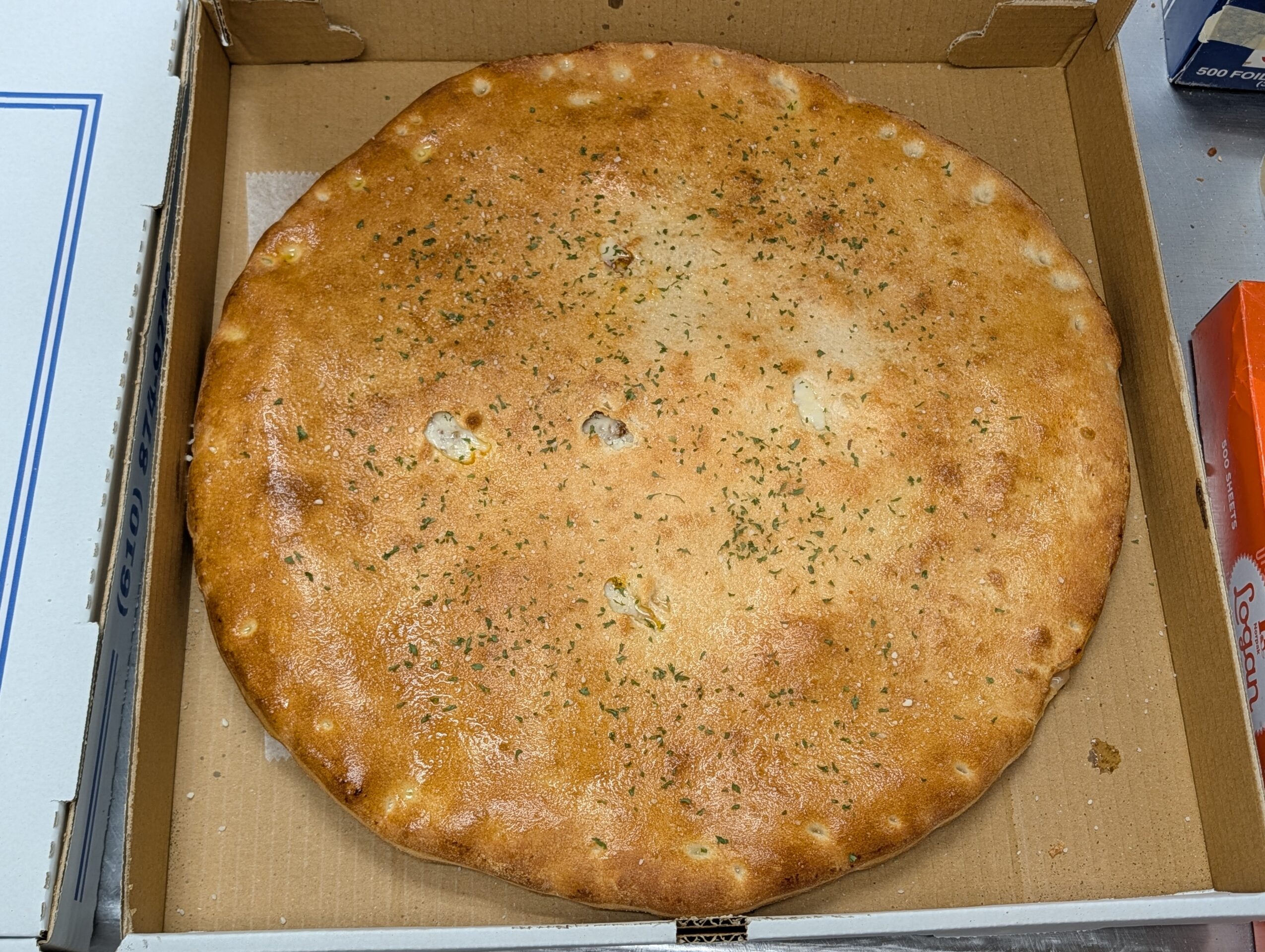 Round, baked flatbread with herbs in a cardboard box, slightly uneven edges, golden-brown surface, and some small holes.