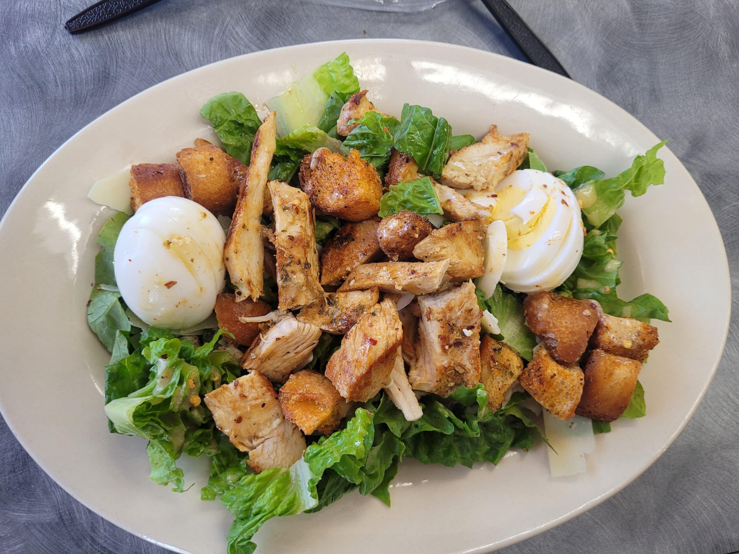 Salad with lettuce, boiled eggs, grilled chicken strips, croutons, and dressing on a white oval plate.