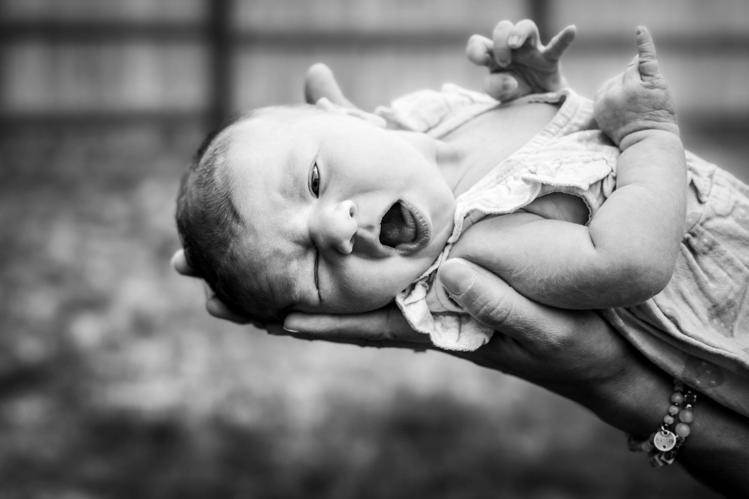 A baby is being held in an adult's hand, yawning with eyes partly closed, outdoors background blurred.