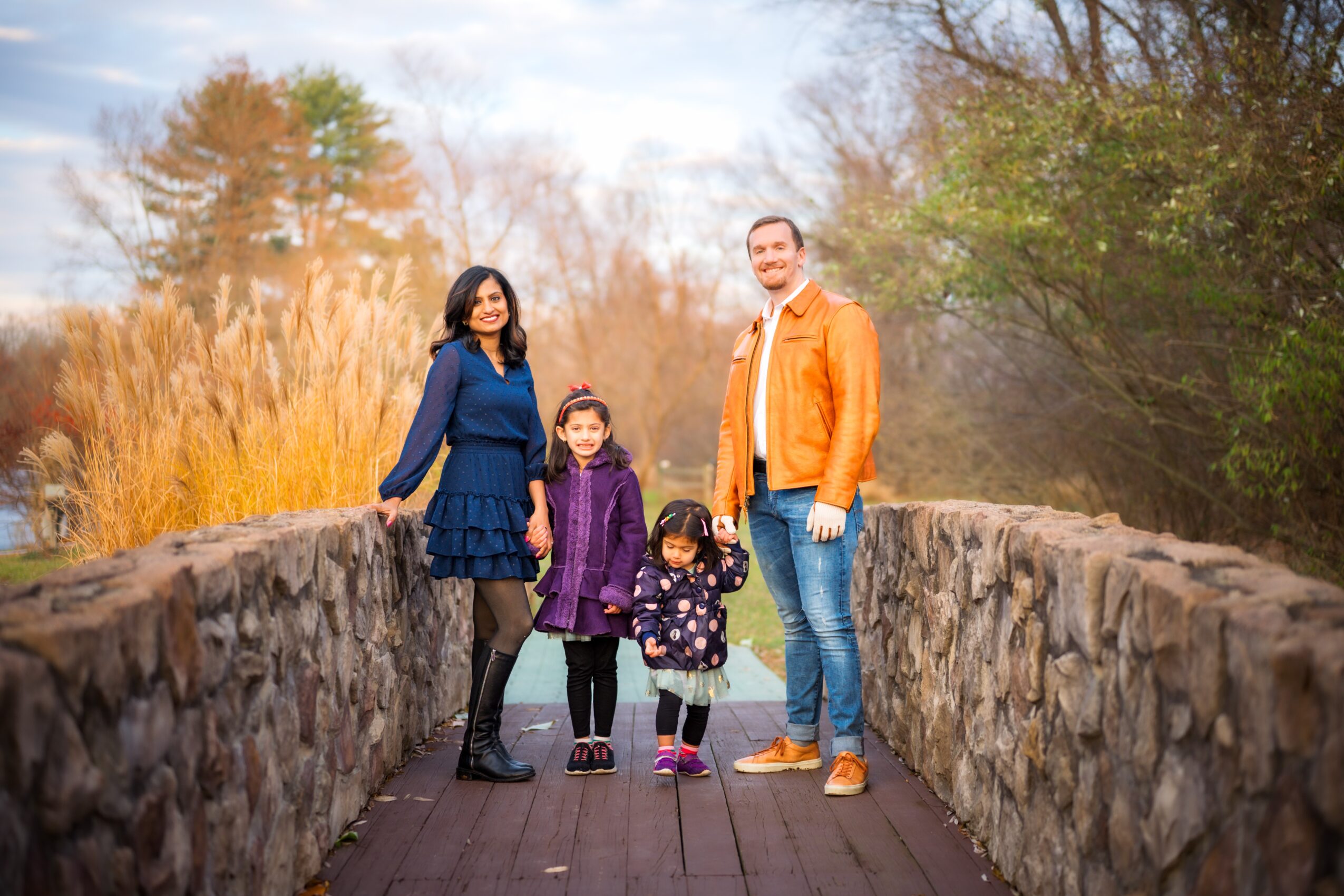 Family of four standing on a stone bridge outdoors, smiling, with trees and sky in background.