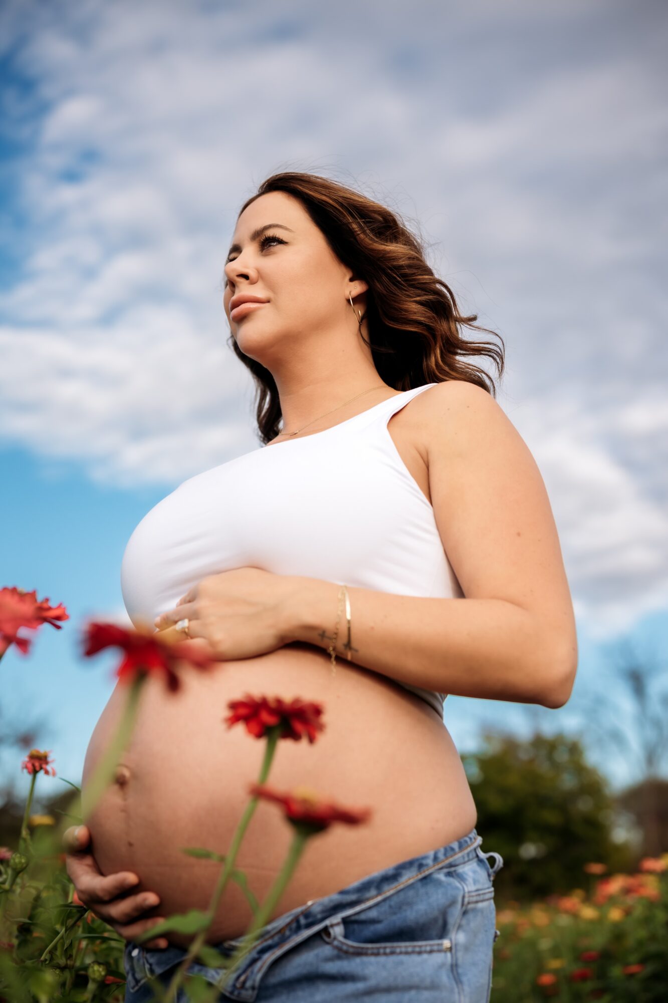 Pregnant woman standing outdoors with sky and clouds, holding her belly, wearing a white top and jeans, flowers in foreground.