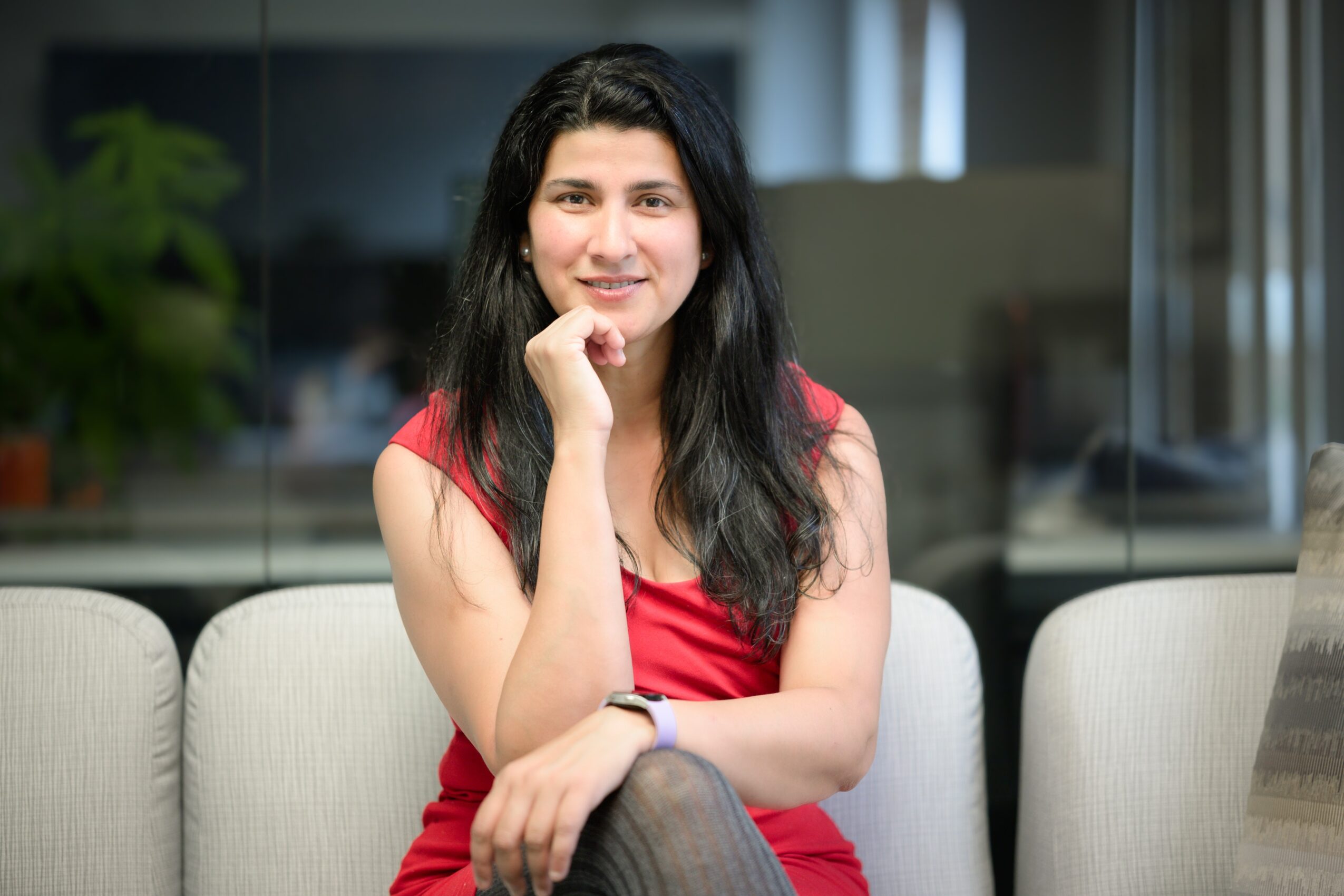 Young woman with long dark hair, smiling, sitting on a light-colored sofa in an indoor setting, wearing a red top.