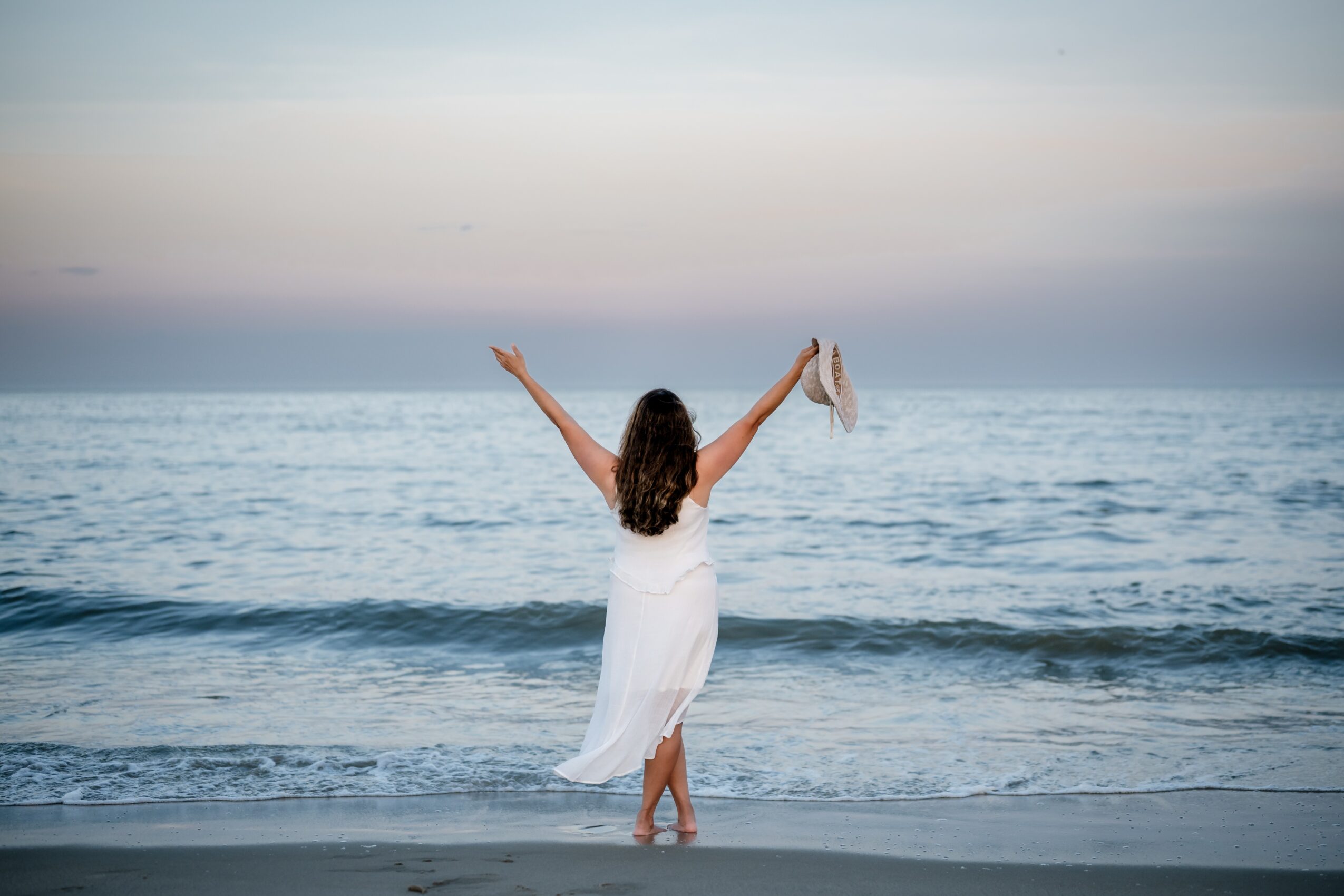 Person in a white dress standing on the beach facing the ocean with arms raised, holding shoes, during sunset or sunrise.