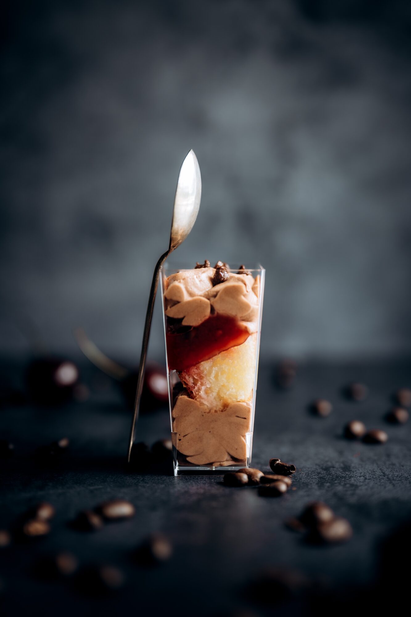 A tall glass of layered dessert with a spoon inserted, surrounded by coffee beans on a dark surface.