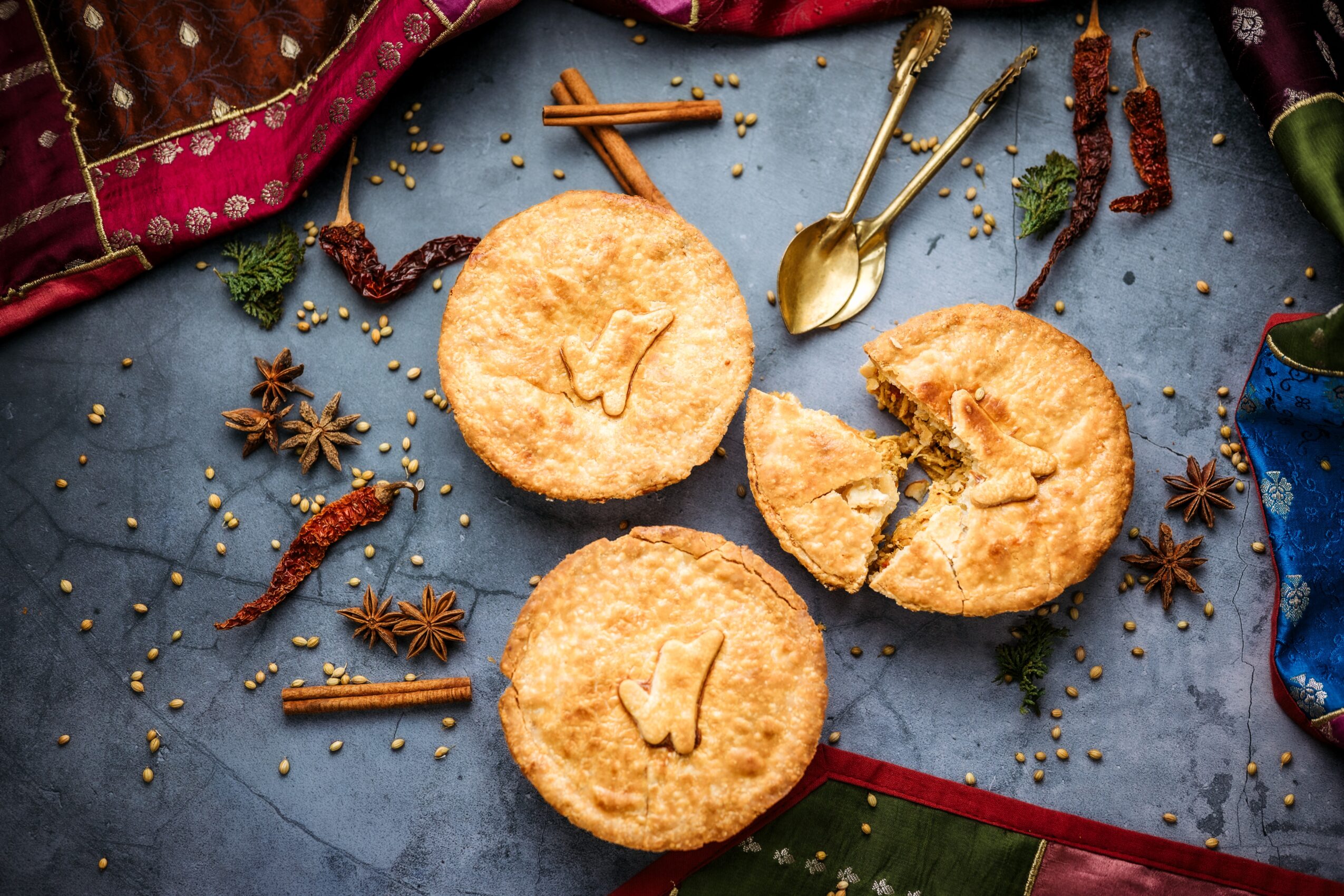 Two round pastries with check marks on top, surrounded by spices, spoons, and colorful fabric on a blue surface.