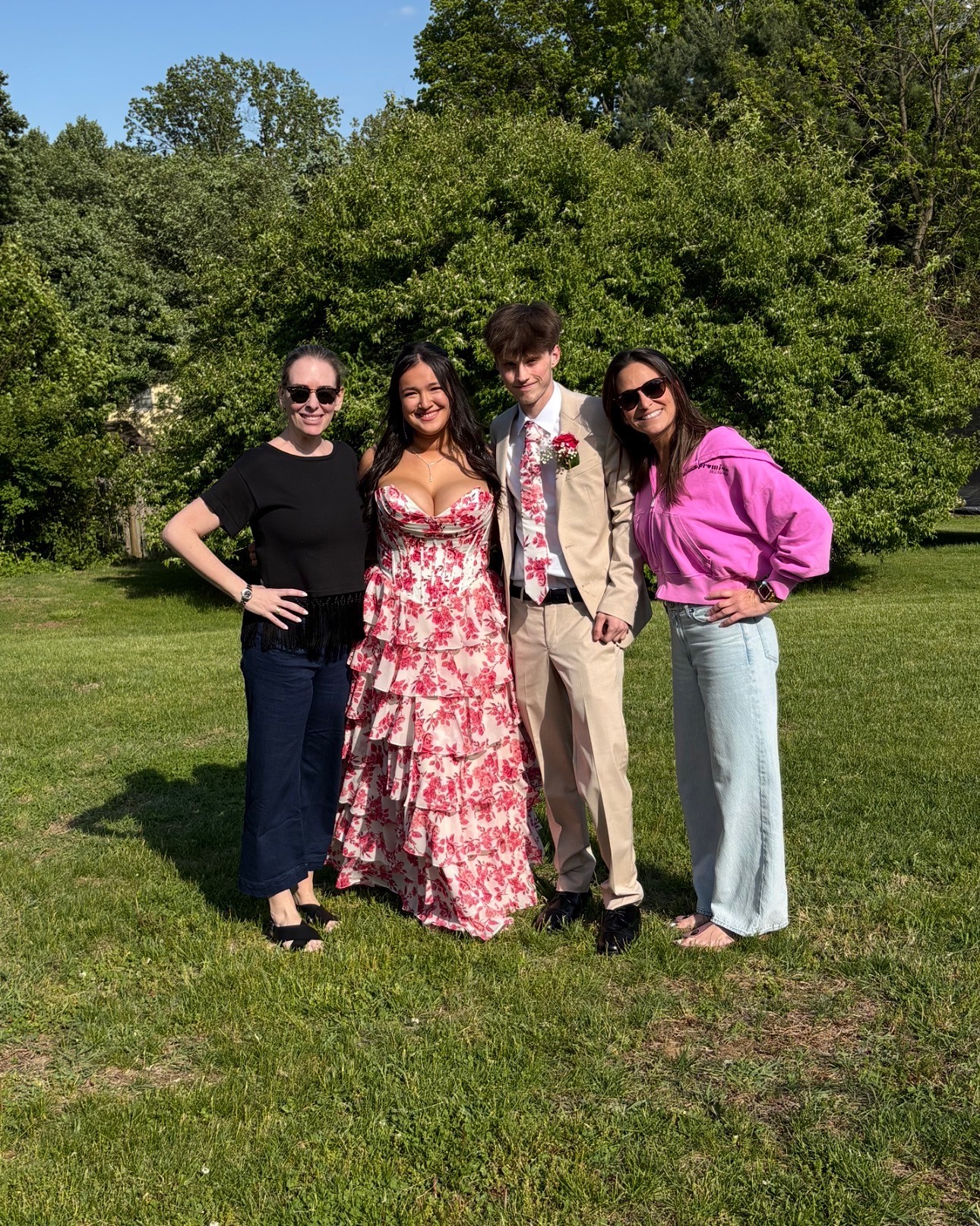 Four people standing outdoors on grass with trees in background, smiling, dressed casually and formally, sunny day.
