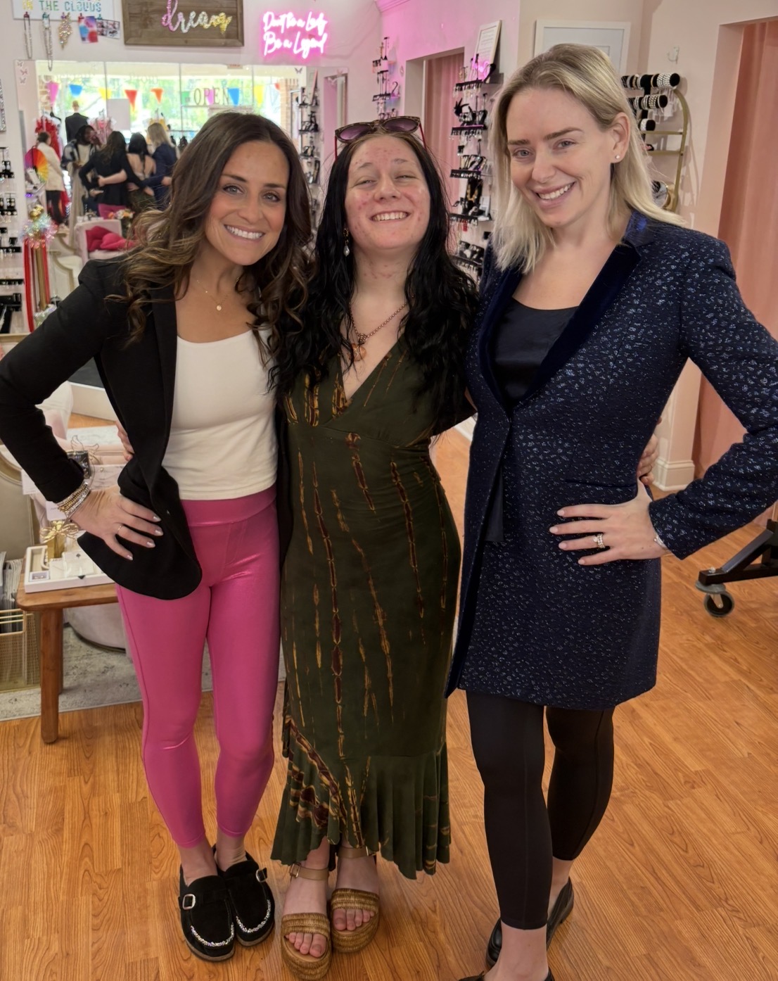 Three women smiling and posing together indoors, with colorful decorations and people in the background.