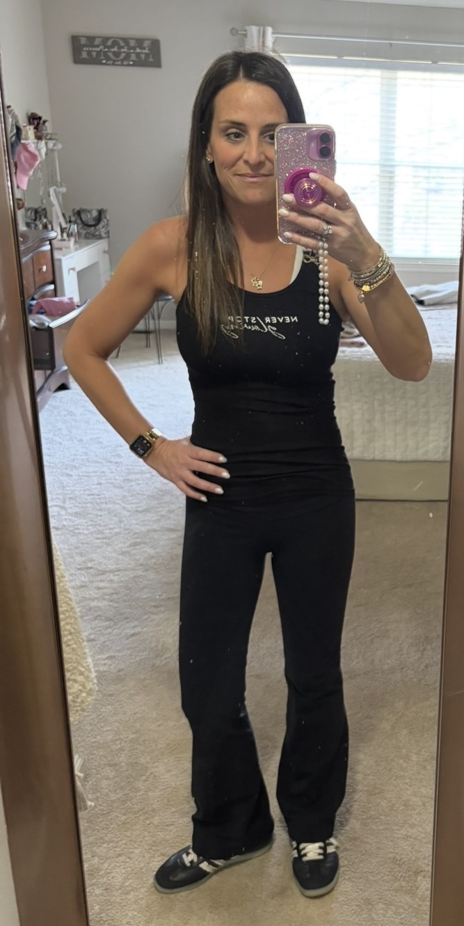 Woman taking a mirror selfie in a room, wearing a black tank top and black pants, holding a phone with a pink case.