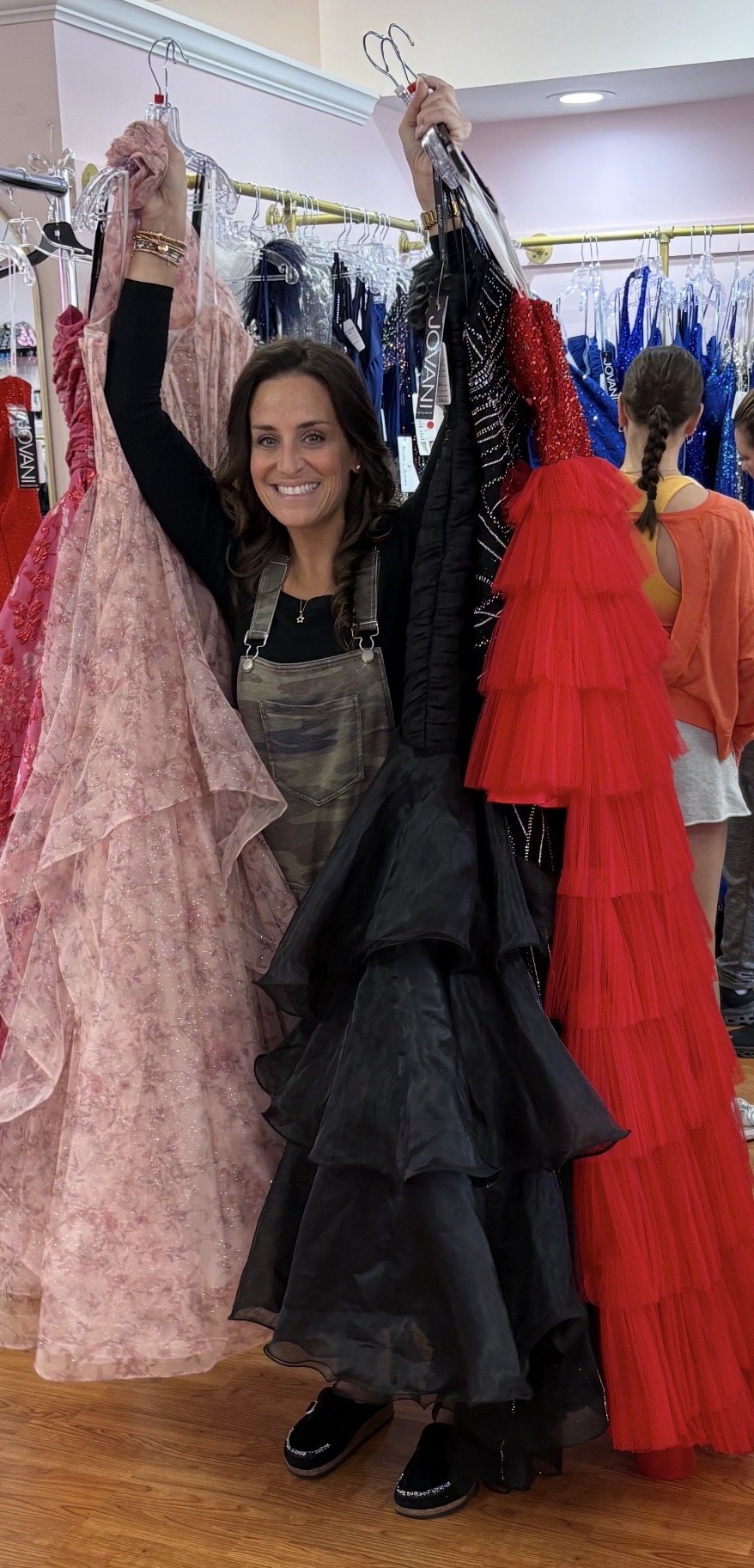 Woman shopping for dresses in a store, holding up pink, black, and red gowns, smiling at camera.