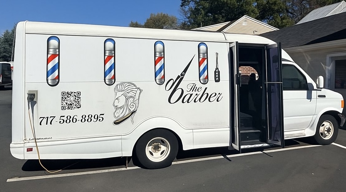 White barber van with barber pole graphics, a lion logo, and contact information, parked on a street.
