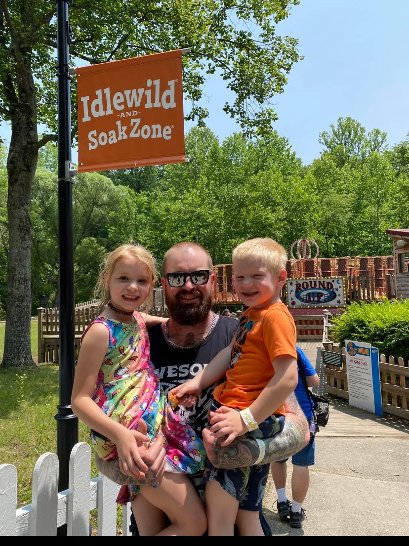 Man with two children, girl in colorful dress and boy in orange shirt, standing outdoors near a sign and fence.