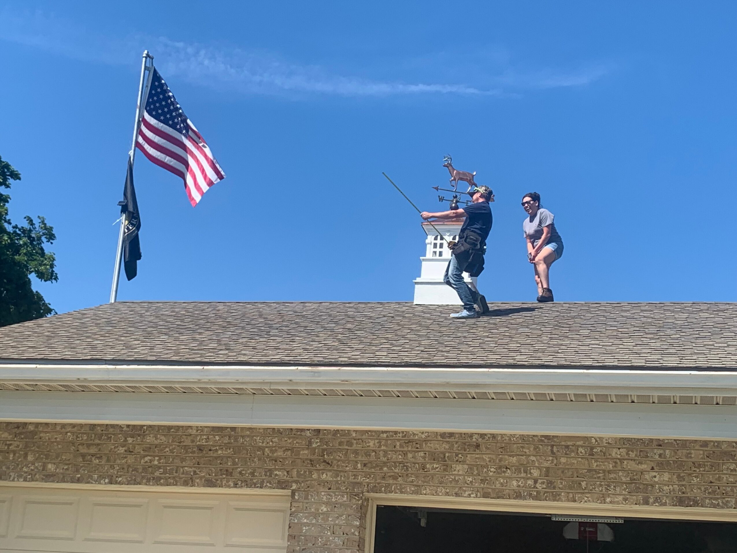 Two people stand on a roof near a flagpole with an American flag, against a blue sky background.