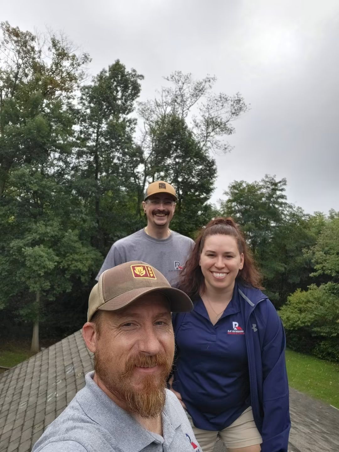 Three people outdoors, two men and one woman, smiling, with trees and cloudy sky in the background.