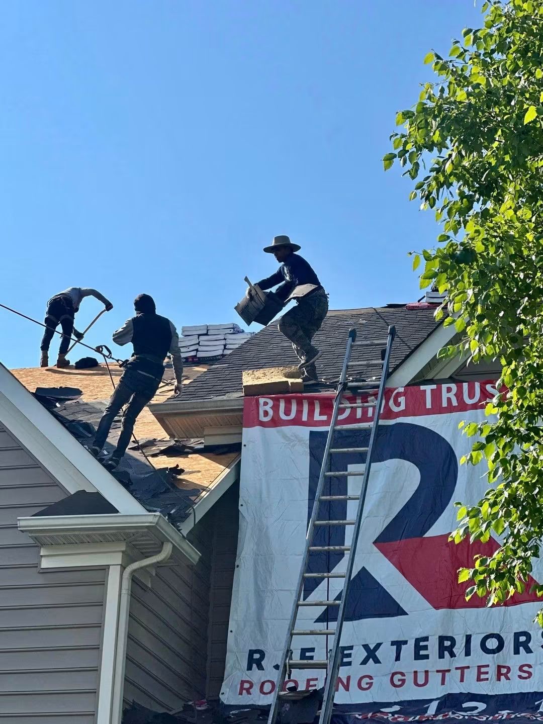 Workers on roof installing gutters, ladder leaning against house, blue sky, tree on right side, building banner visible.