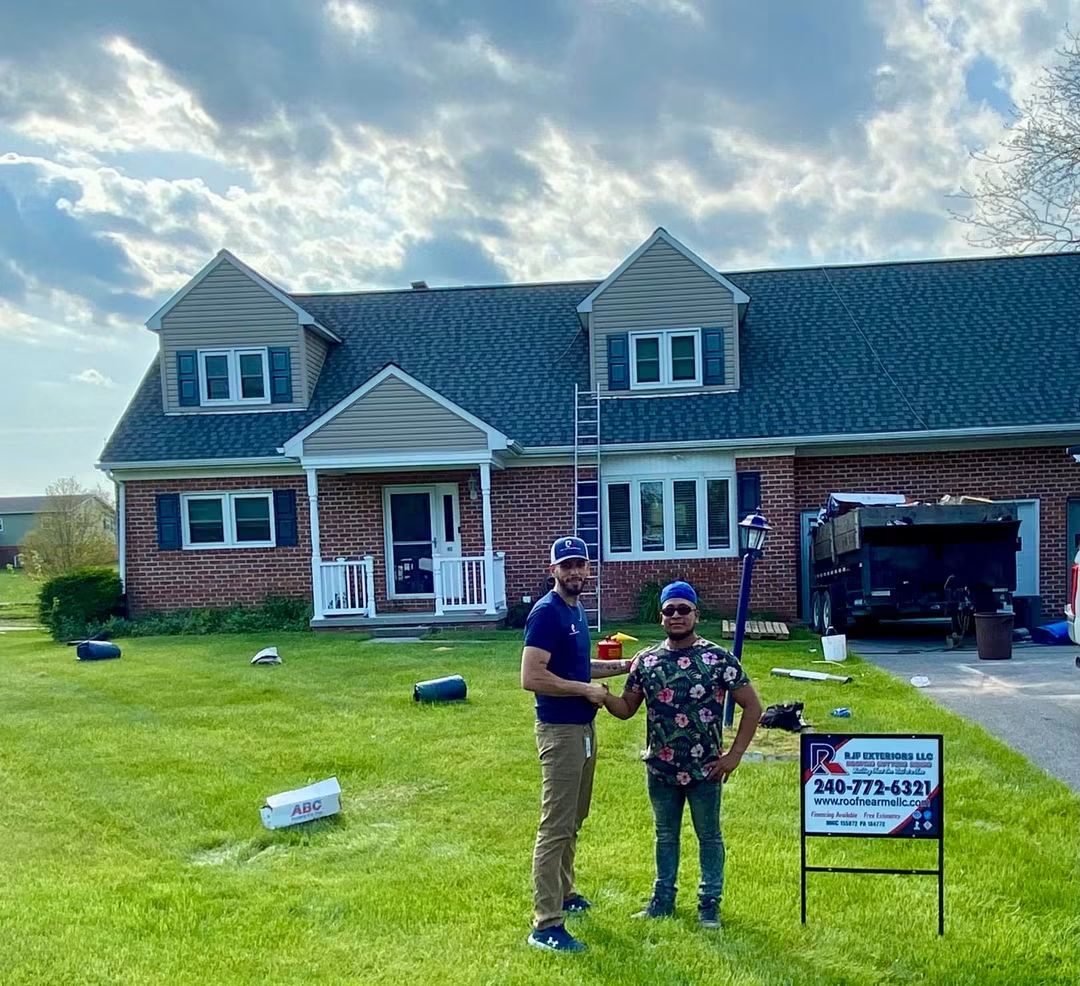 Two people stand on a lawn in front of a large brick house with dormer windows, a sign, and a truck. Cloudy sky.