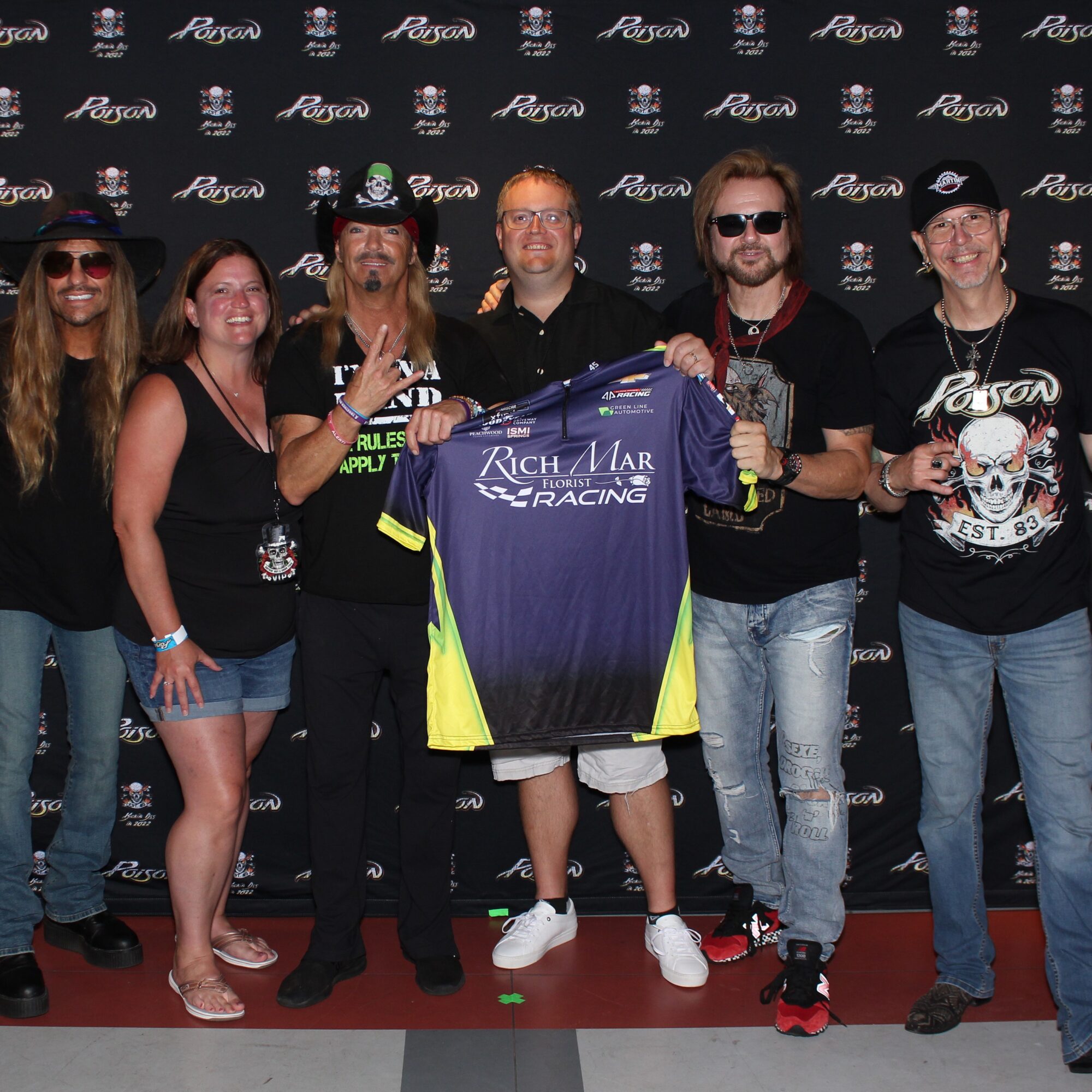 Group of six people standing together, holding a racing jersey, in front of a branded backdrop, smiling and celebrating.