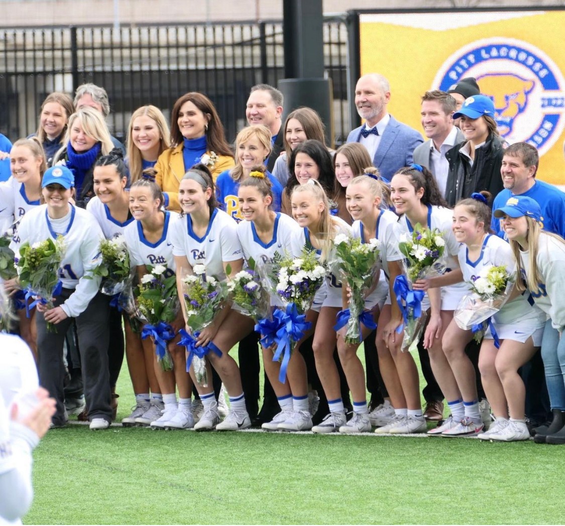Group of young female athletes in sports uniforms holding flowers, with adults behind them, on a sports field.