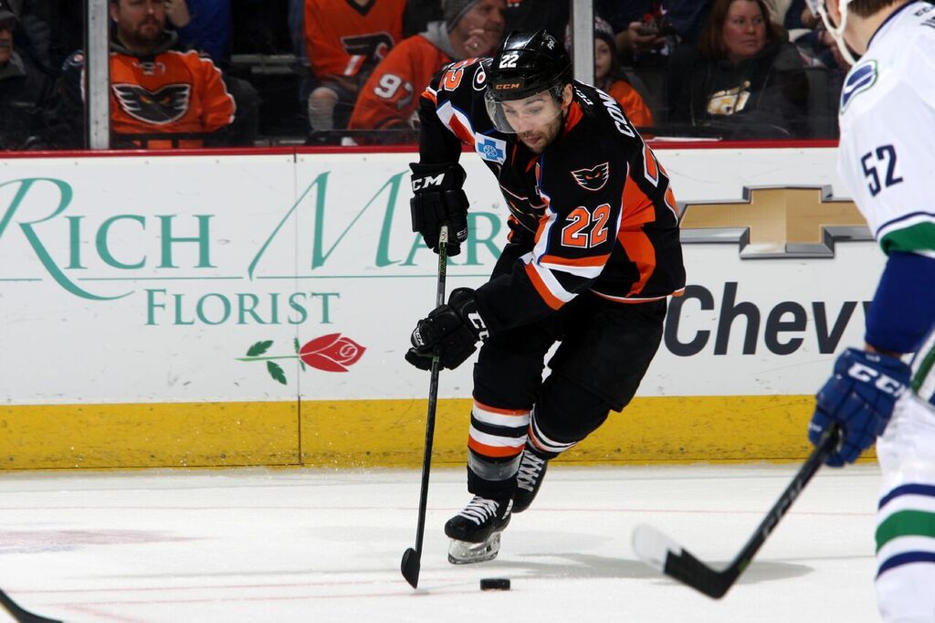 Hockey player in black and orange uniform skating on ice during game, with spectators in background.