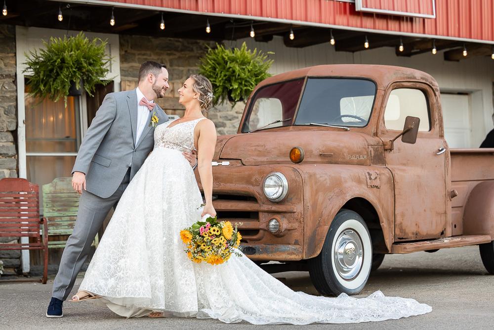 Bride and groom in wedding attire standing next to a rusty vintage truck outside a building with plants.