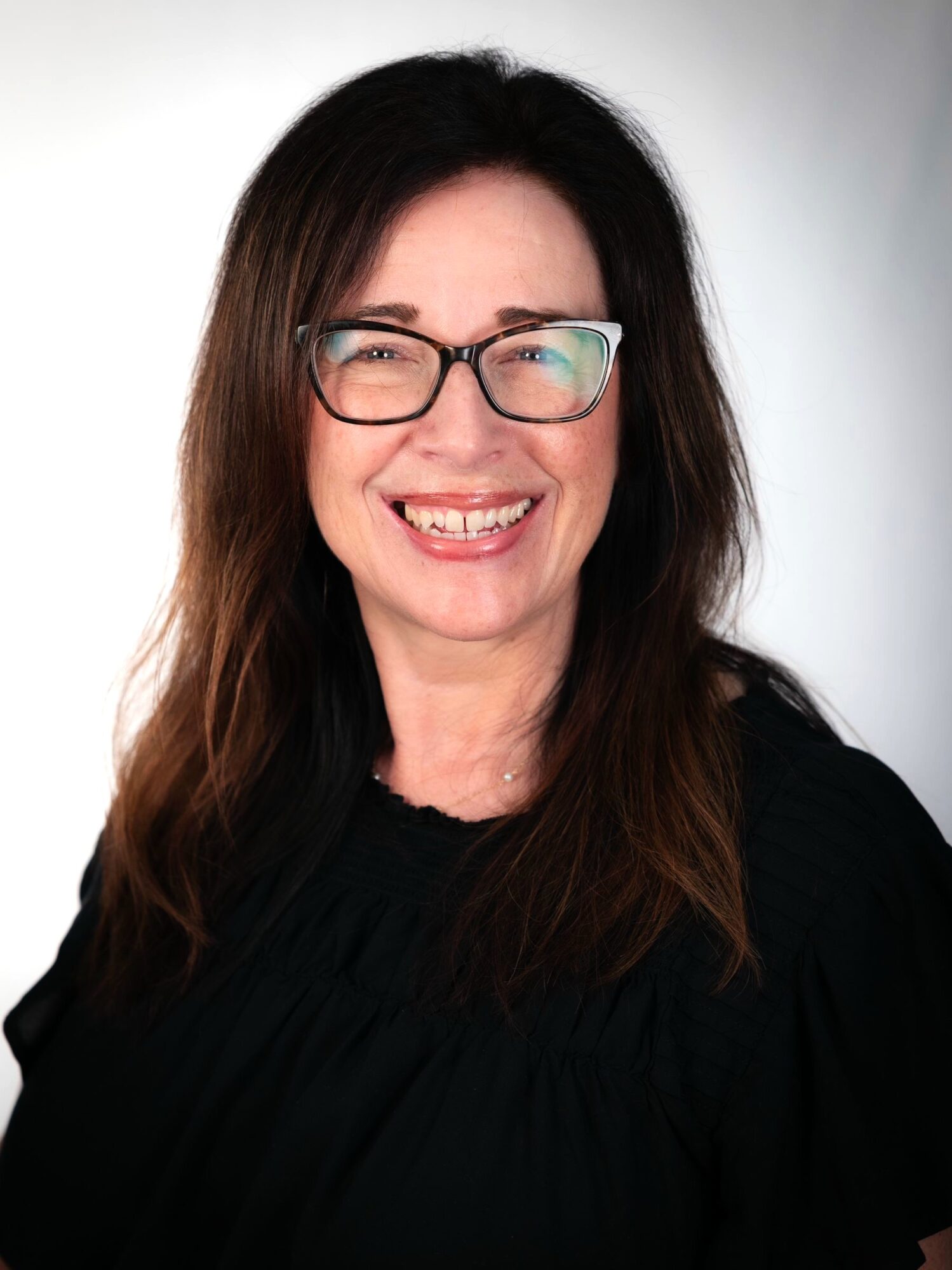 Woman with long dark hair, glasses, smiling, wearing a black top, against a light background.