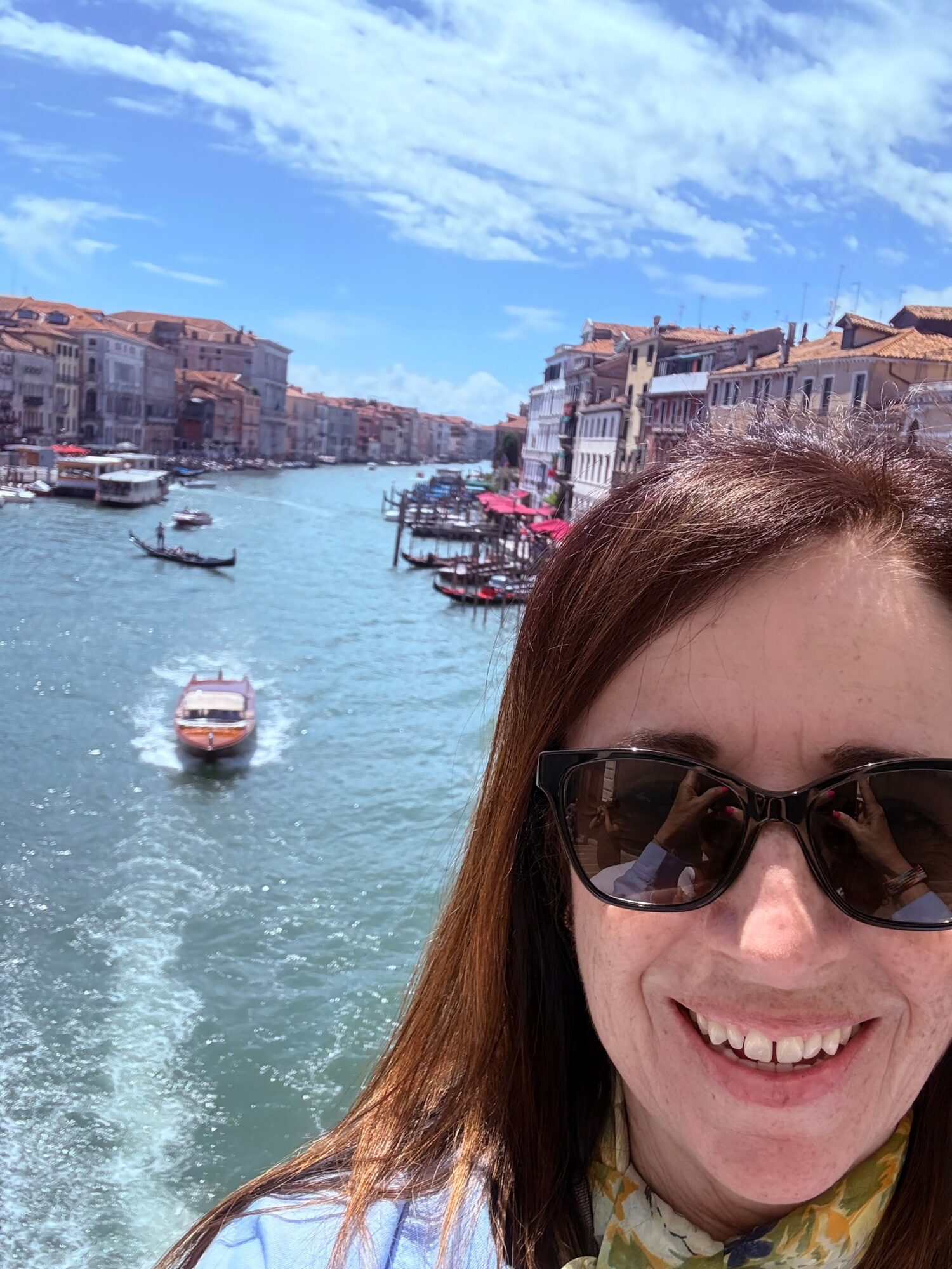Smiling woman wearing sunglasses takes a selfie with a canal, boats, and colorful buildings under a blue sky in the background.