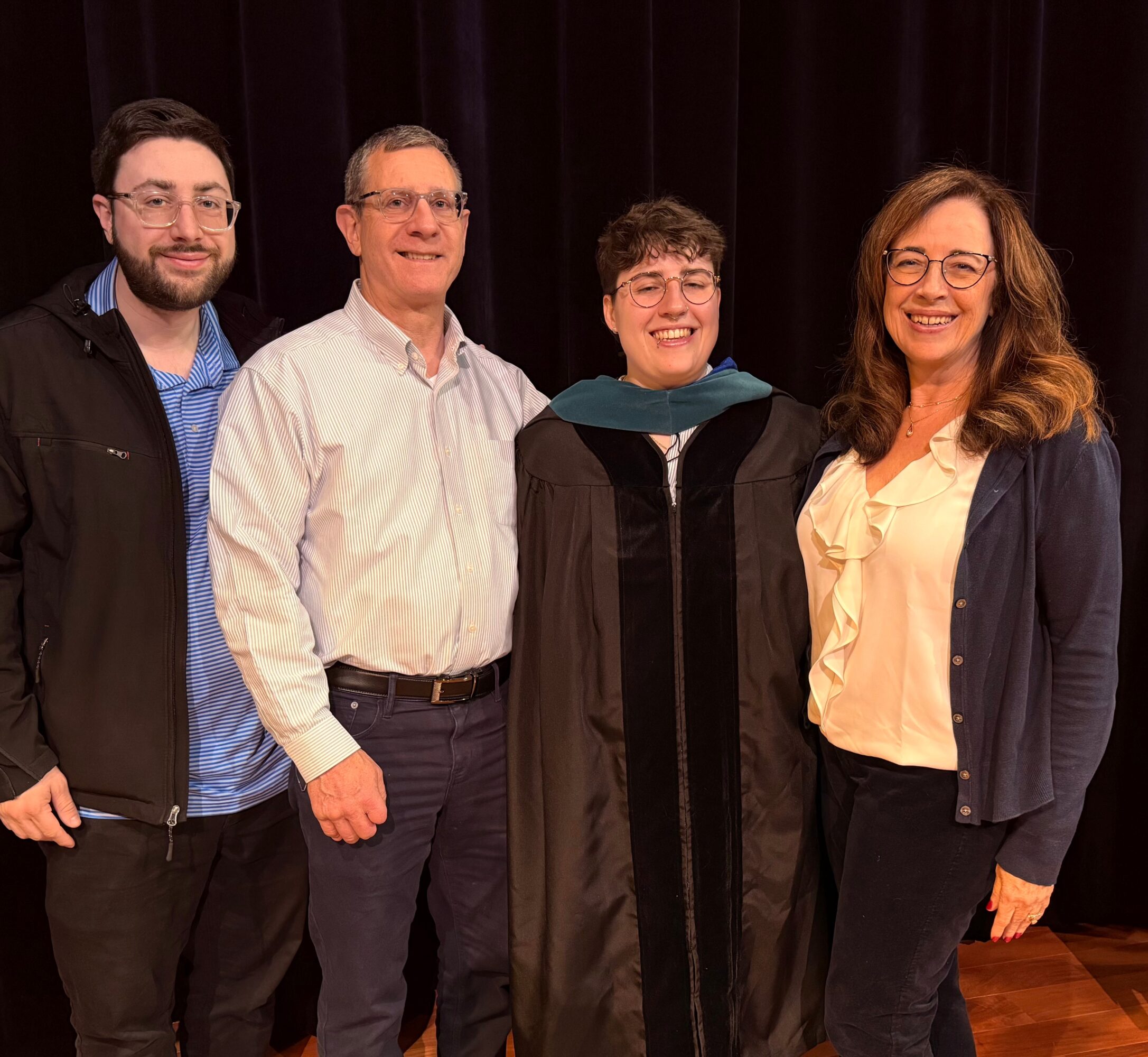 Four people standing together indoors, smiling, with a dark curtain background.