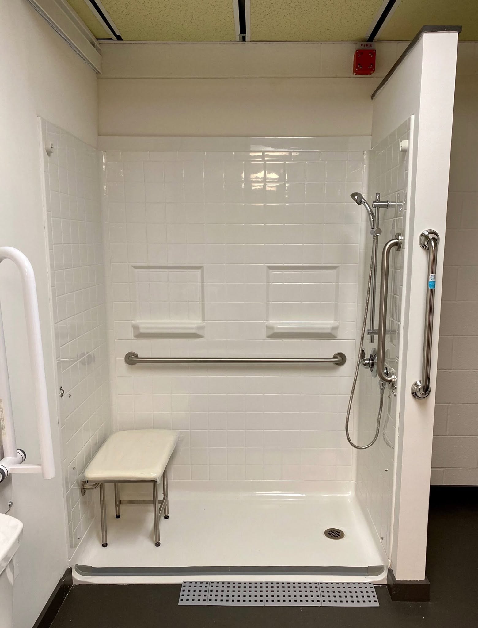 Accessible shower area with a bench, grab bars, and a handheld showerhead, enclosed by a partial wall, with a drain on the floor.