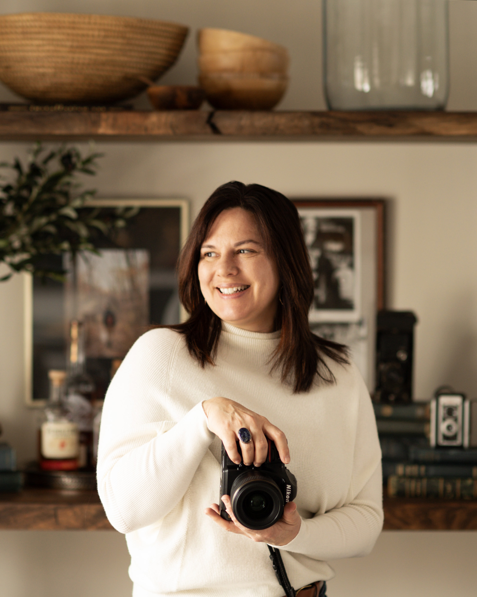 Woman smiling and holding a camera indoors, with framed pictures and decor in the background.