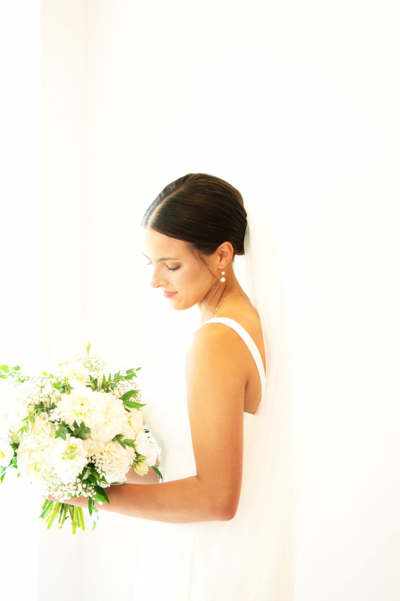 Woman in white dress holding a bouquet of white flowers, looking down, with dark hair styled in an updo, standing against a white background.