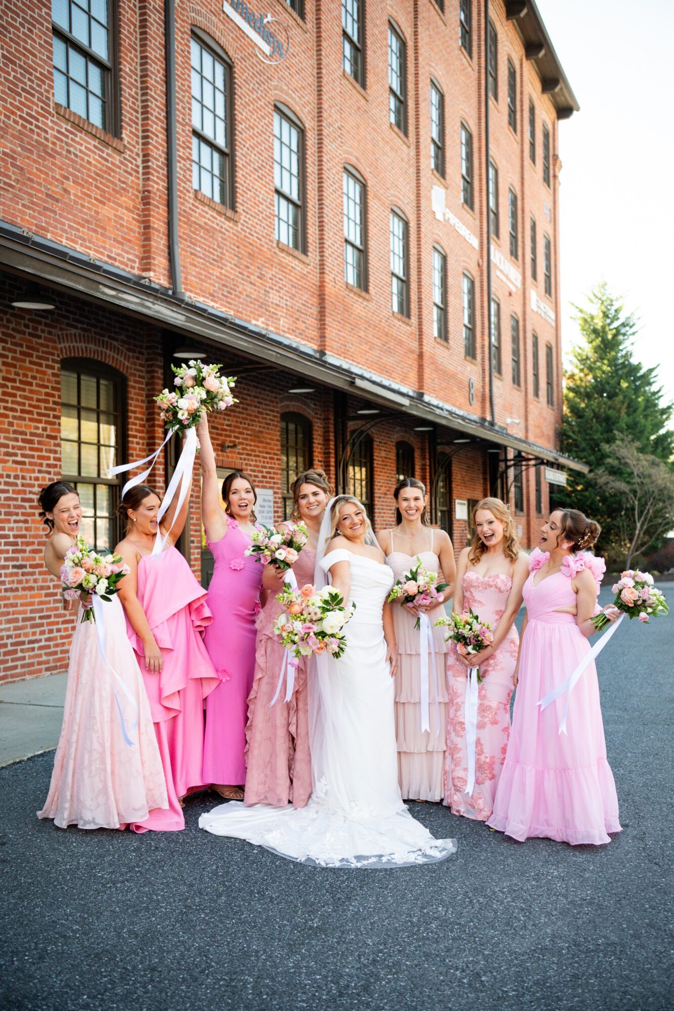 Group of women in pink and white dresses holding bouquets, standing outside a brick building, celebrating a wedding.