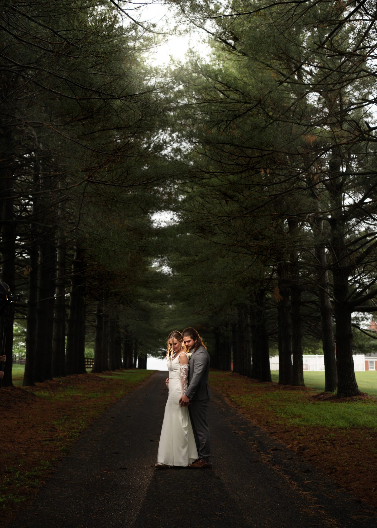 A couple in wedding attire standing on a forest path, surrounded by tall trees, with sunlight filtering through the branches.