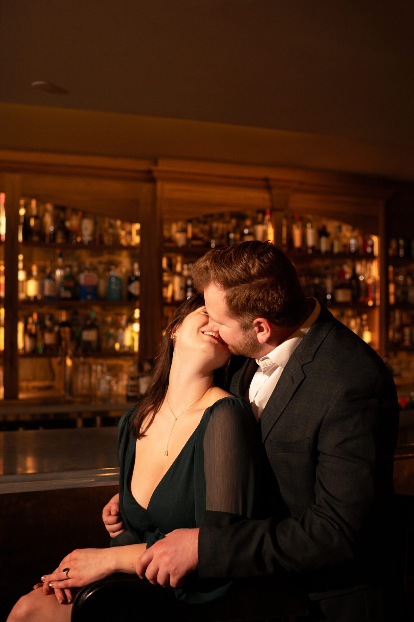 A couple kissing in a bar with shelves of bottles in the background.