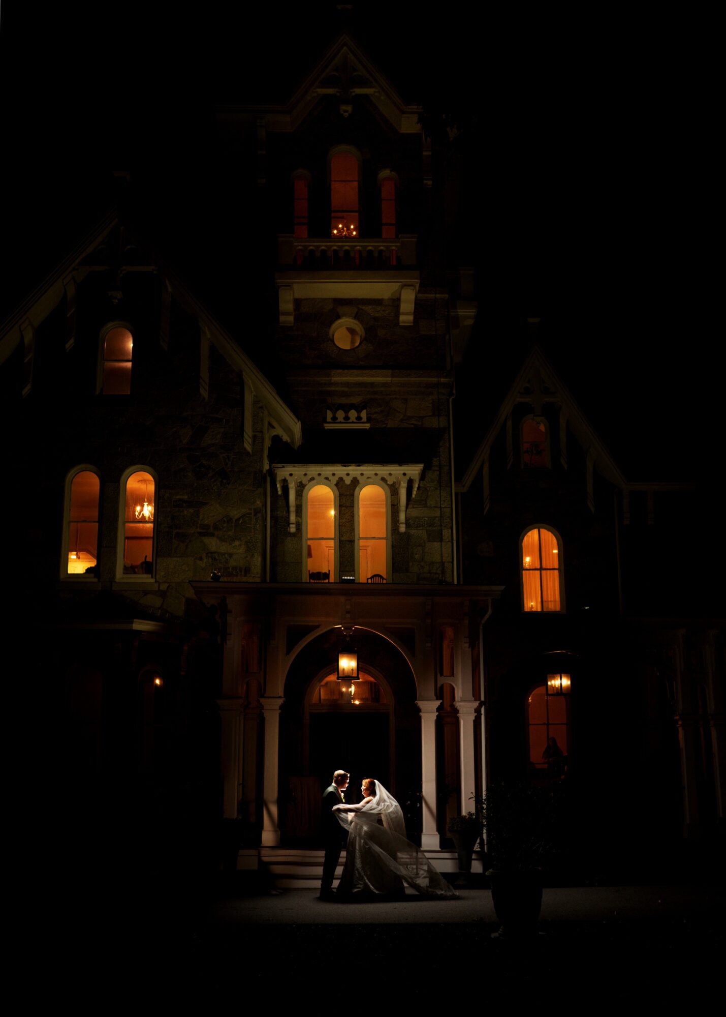 A couple dancing on a porch in front of a large, illuminated Victorian-style house at night.