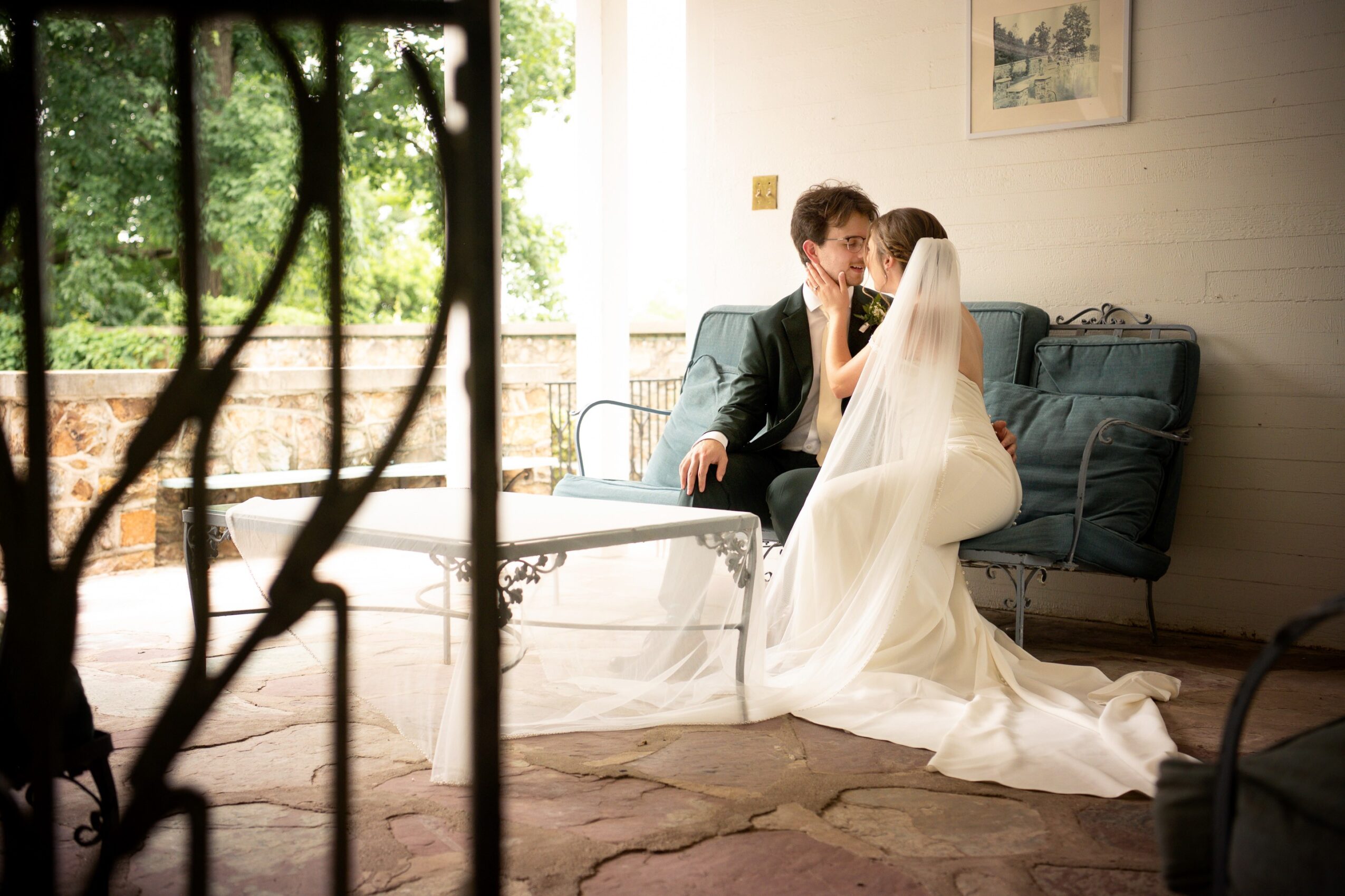 A bride and groom sit closely on a sofa indoors, with a window showing greenery outside, during a wedding moment.