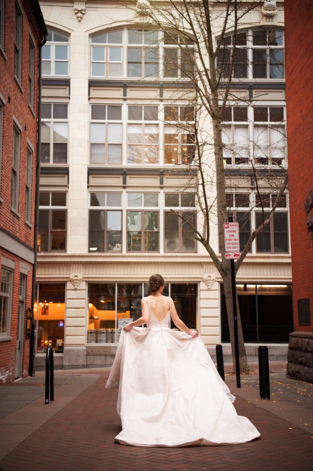 Woman in wedding dress standing on city sidewalk with tall building and tree behind her.