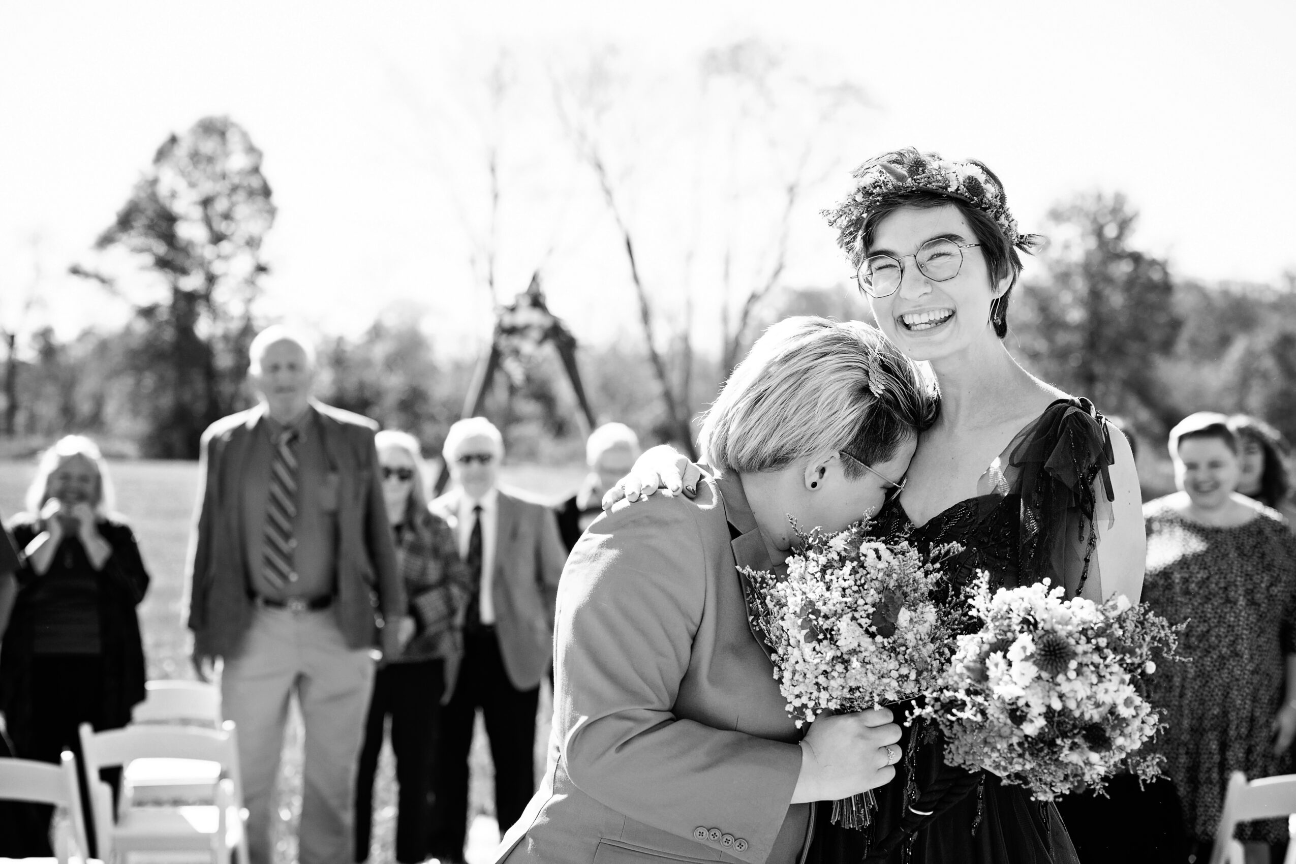 Two women embrace, one holding a bouquet of flowers, smiling, with a group of people outdoors in the background.