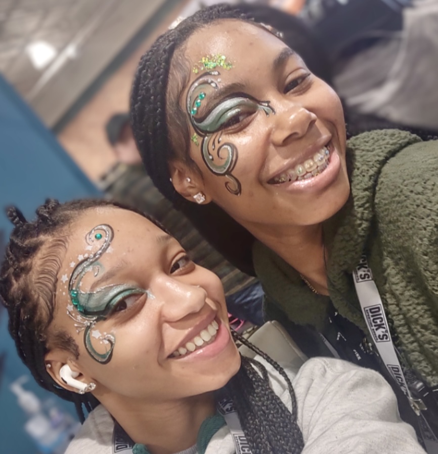 Two smiling women with face paint and braided hair, close together, indoors. One wears a green jacket, the other a gray top.