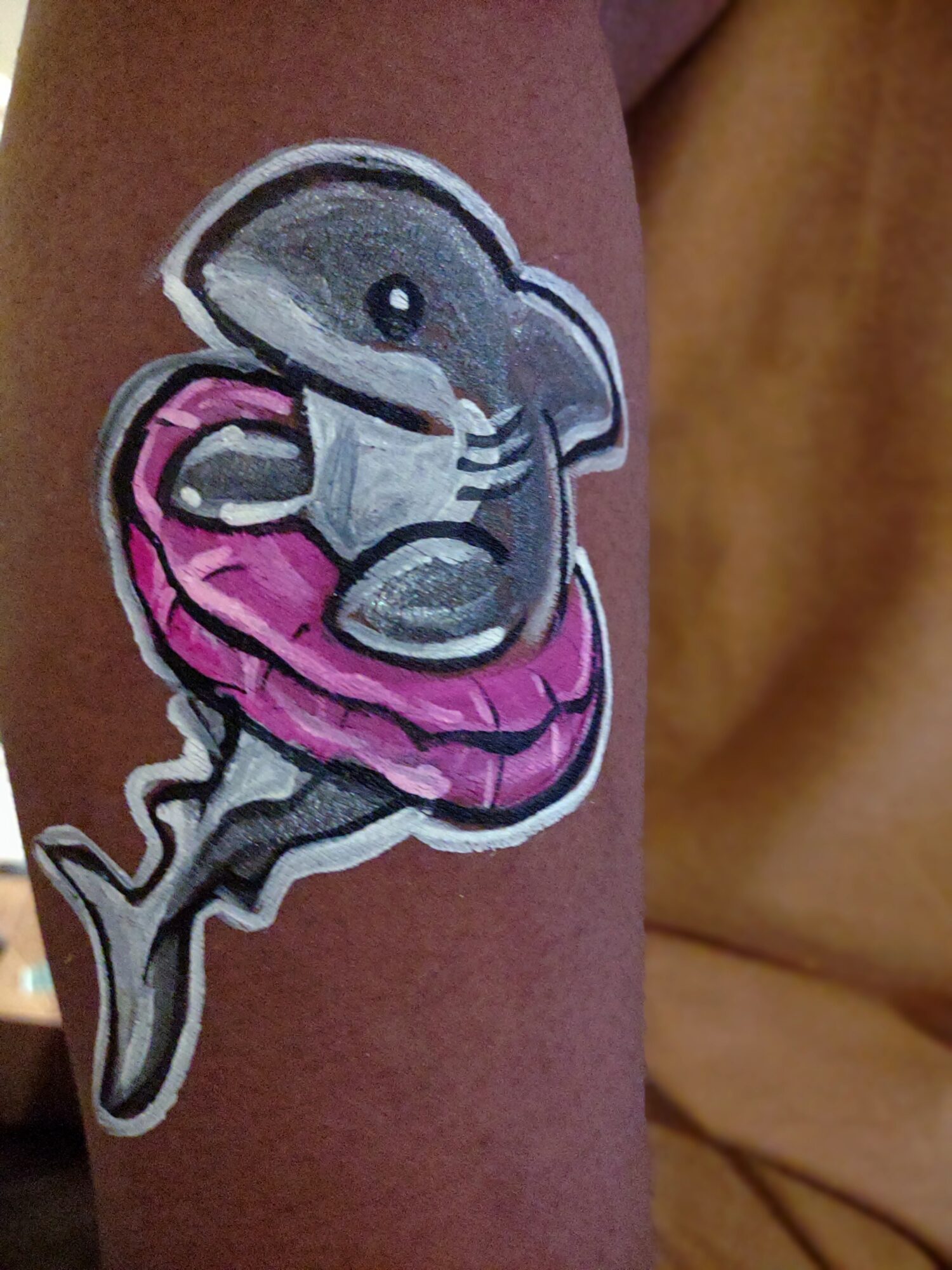 Tattoo of a cute dolphin wearing a pink inflatable ring, holding a small fish, on skin.
