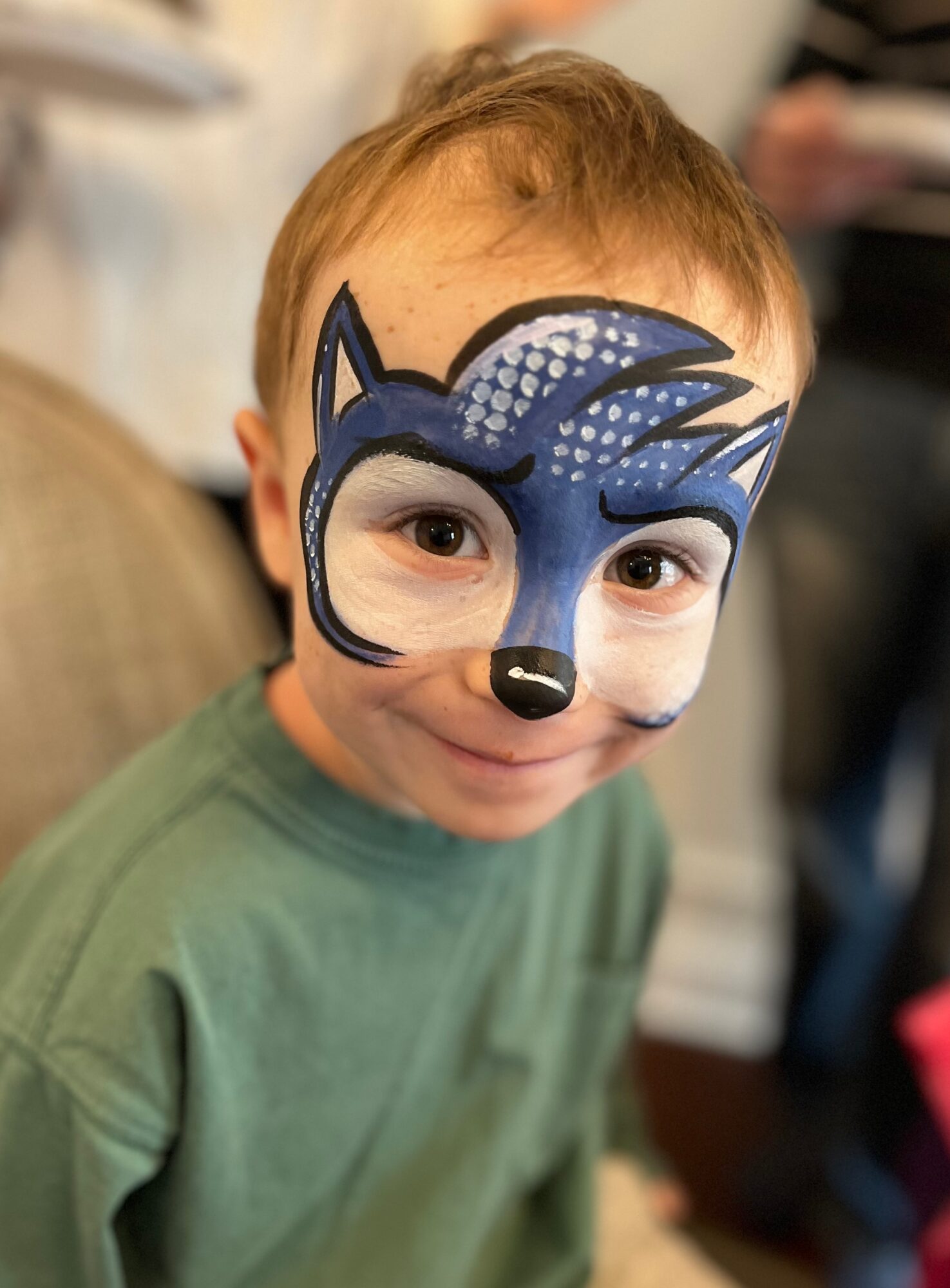 Child with a blue and white animal face paint mask, smiling, wearing a green shirt, indoors.