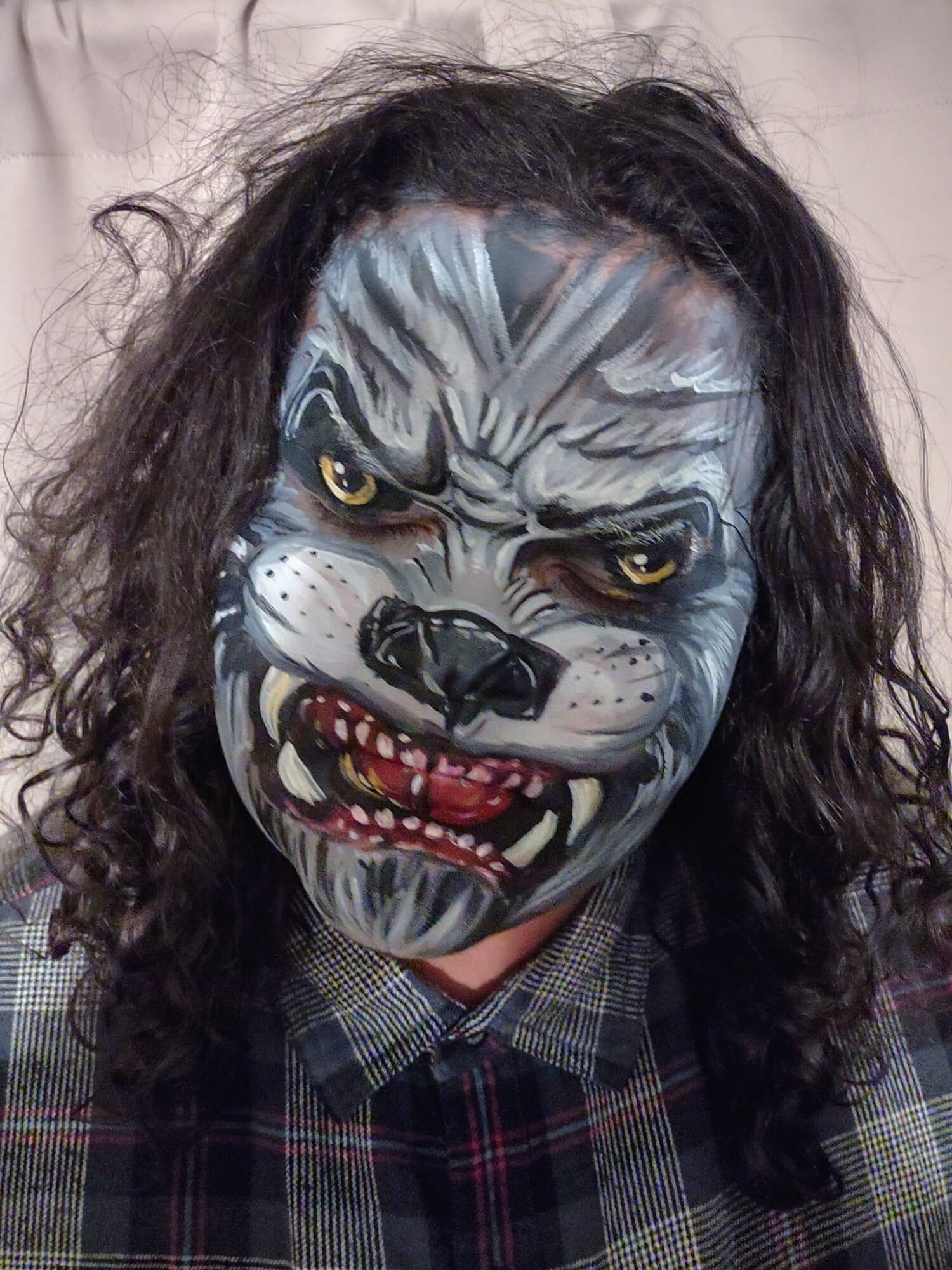 Person with detailed tiger face paint, snarling with teeth showing, long curly hair, wearing a checkered shirt.