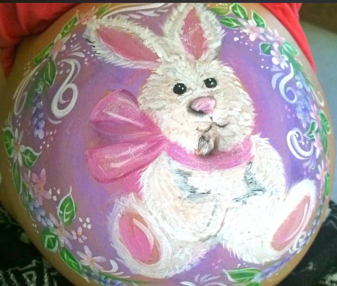 Decorative painted egg with a white bunny wearing a pink bow, surrounded by floral patterns on a purple background.