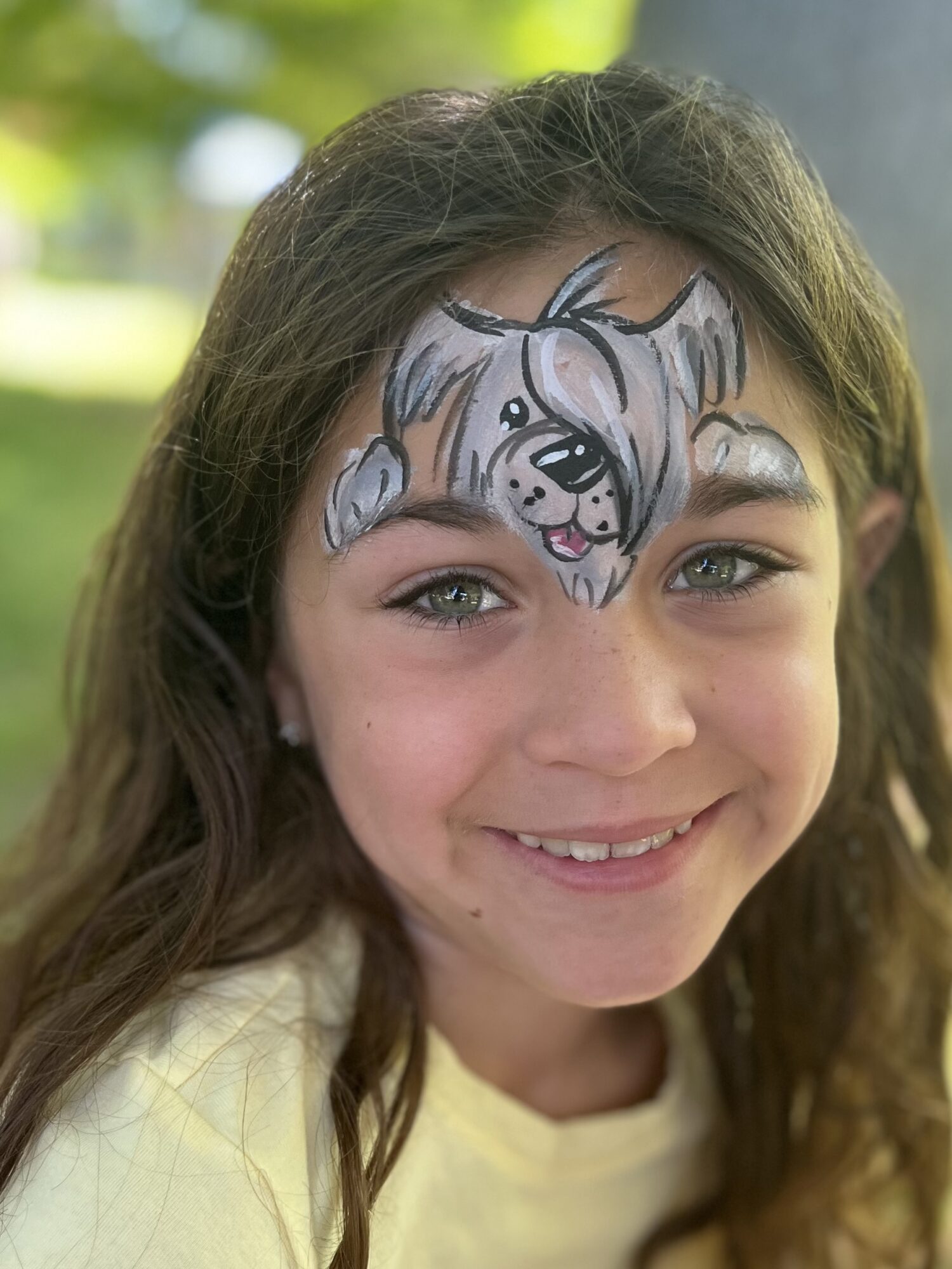 Young girl with face paint of a cartoon dog on her forehead, smiling outdoors with blurred greenery in background.