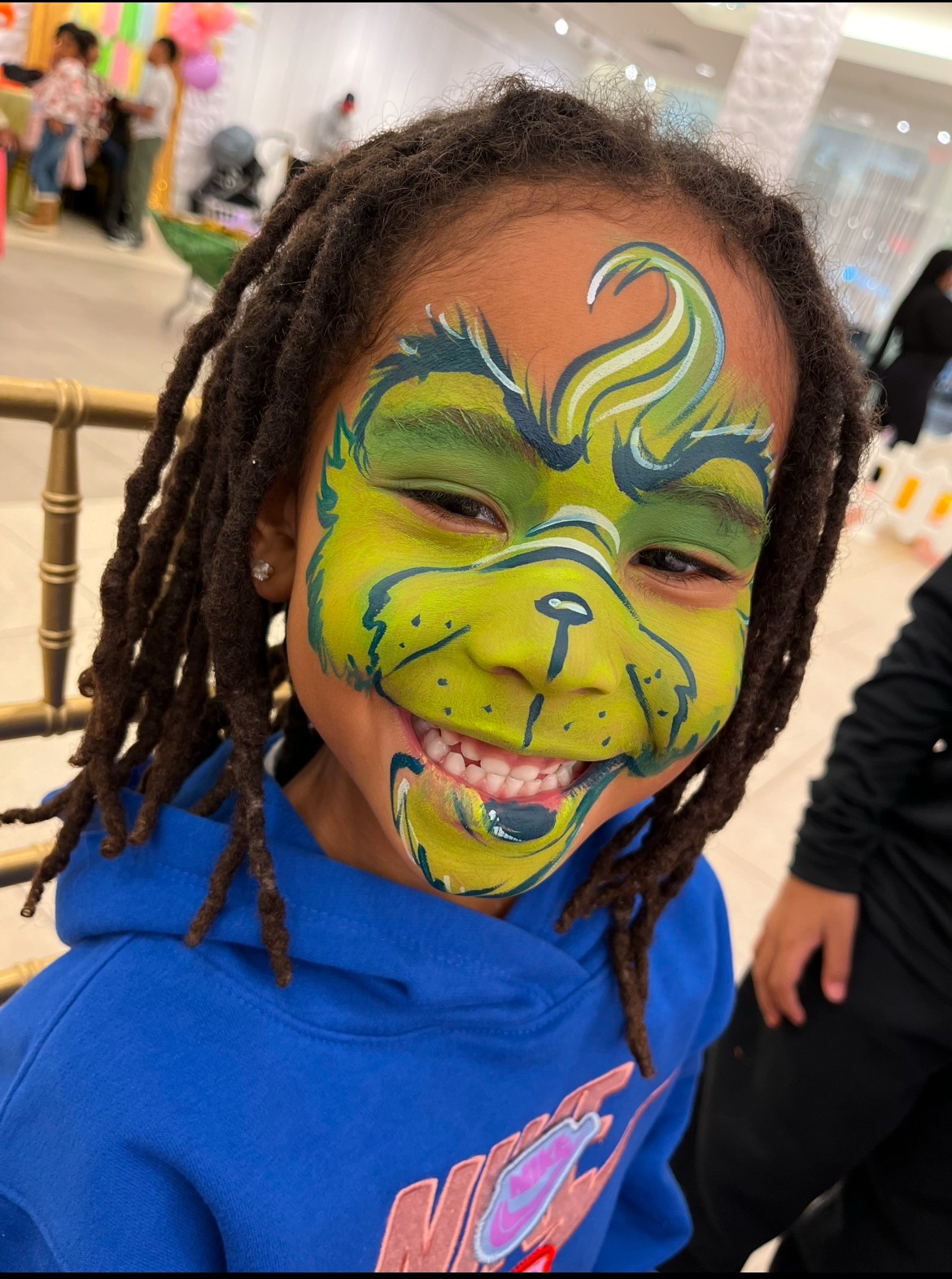 Child with face paint of a colorful dog, smiling, wearing a blue hoodie, in a busy indoor setting.