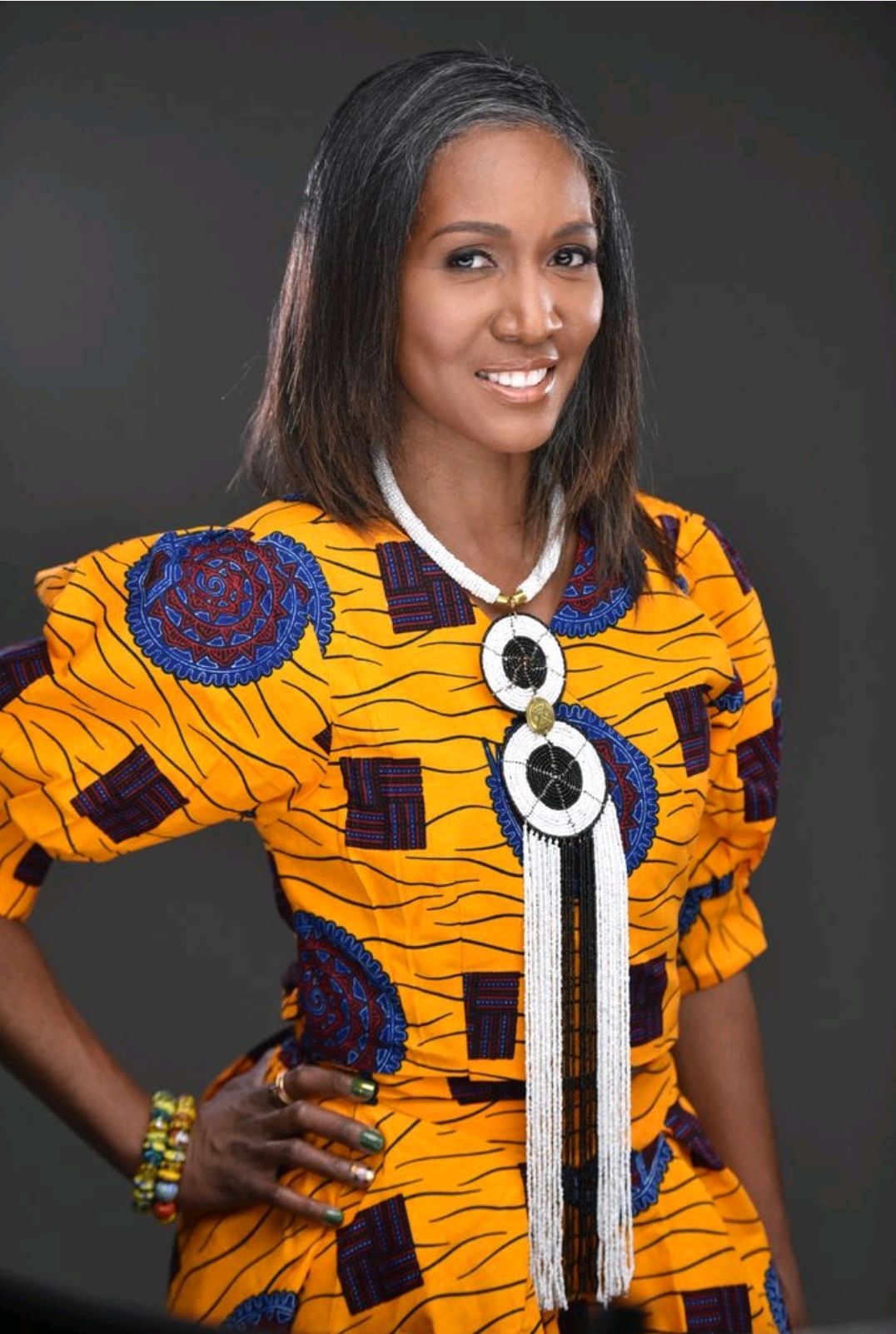 Woman with shoulder-length hair wearing a colorful patterned dress, smiling, against a dark background.